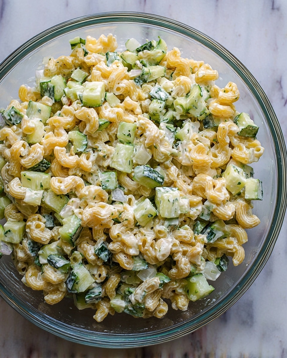 A clear glass bowl filled with a creamy pasta salad is shown from above. The salad has two main layers mixed together: short, curved yellow pasta pieces with a textured surface, and chopped cucumber pieces that are green on the skin and pale inside. Small bits of white onion and specks of herbs are scattered throughout. The creamy dressing coats all ingredients with a slightly shiny look. The bowl is placed on a white marbled surface. photo taken with an iphone --ar 4:5 --v 7