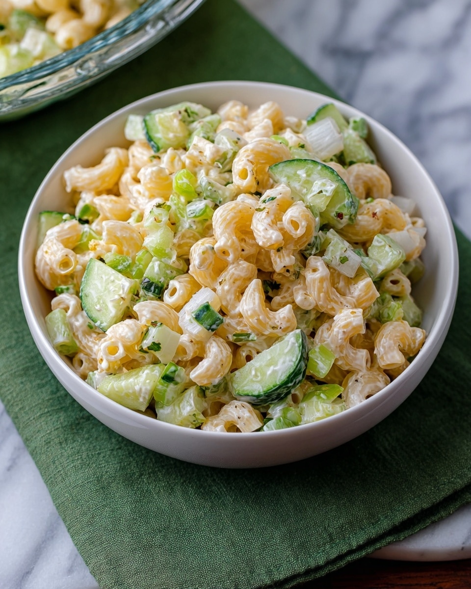 A white bowl filled with a creamy macaroni salad, showing three main layers: the pale yellow curved macaroni pasta, scattered with bright green cucumber slices and celery chunks, and small white onion pieces mixed all together. The salad is coated in a light creamy dressing that gives a soft shine to the pasta and vegetables. The bowl is placed on a green cloth, set on a white marbled surface with part of a glass bowl of the same salad visible at the bottom edge. Photo taken with an iphone --ar 4:5 --v 7