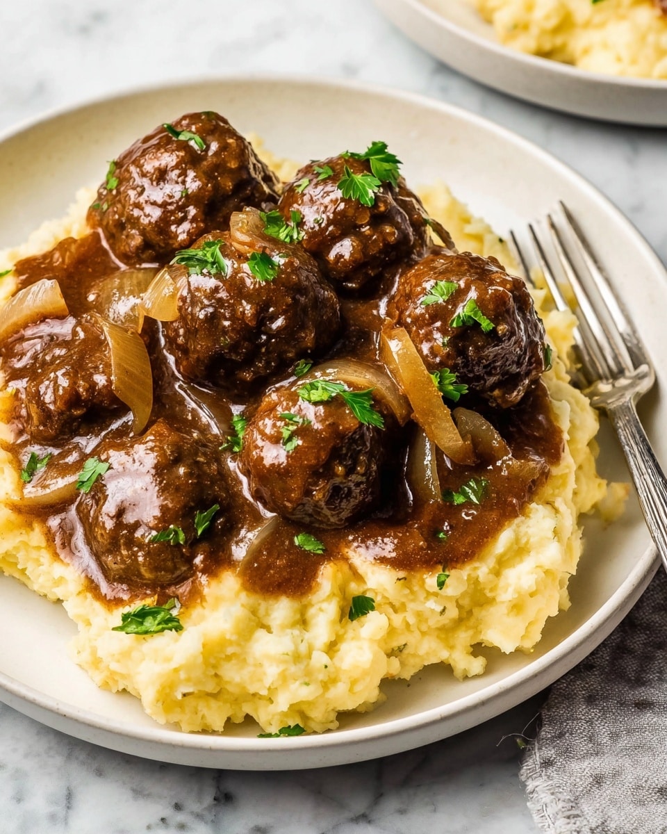 A white plate holds a serving of mashed potatoes topped with a rich brown sauce filled with several meatballs and cooked onion slices. The mashed potatoes form the bottom layer, soft and light yellow in color with a slightly chunky texture. On top, the thick brown sauce coats the meatballs and onions, giving them a glossy shine. The meatballs are round and dark brown with a slightly rough surface. Small green parsley leaves are sprinkled over the meatballs and sauce for color contrast. The plate sits on a white marbled surface with a silver fork beside it. photo taken with an iphone --ar 4:5 --v 7