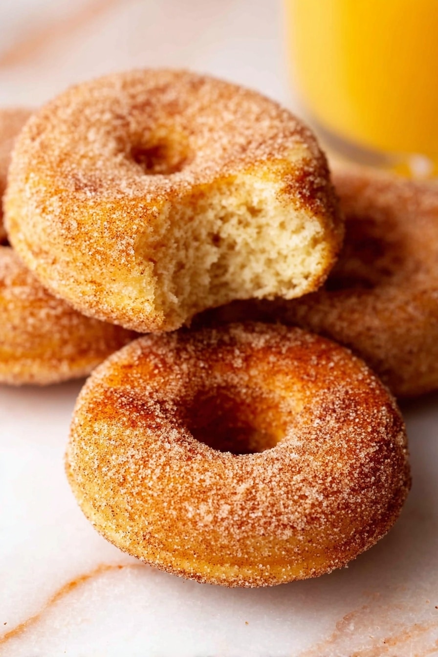 Vegan Apple Cider Donuts, vegan fall donuts, vegan apple cider treats, easy vegan donut recipe, cozy fall dessert - The image shows three cinnamon sugar donuts with a light golden brown color and a textured coating of sugar and cinnamon. One donut is in the front with a bite taken out, revealing a soft, fluffy inside with a slightly crumbly texture. The other two donuts are whole and placed closely behind it on a light-colored surface. The background has a white marbled texture, and there is a glass of orange juice visible on the right side of the image. photo taken with an iphone --ar 2:3 --v 7