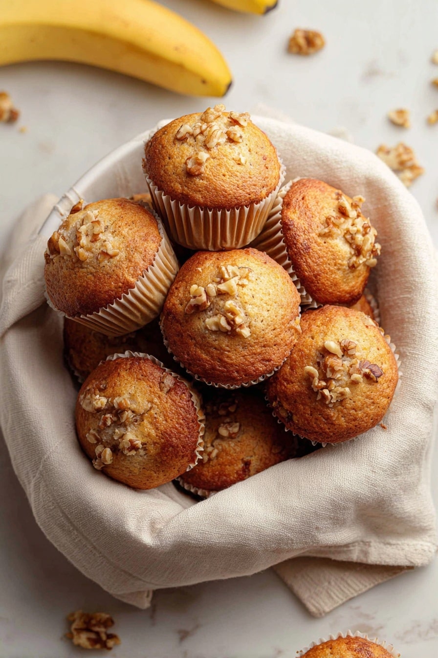 Quick & Easy Vegan Banana Muffins, vegan banana muffins, healthy vegan muffins, fluffy banana muffins, quick vegan baking - A white bowl lined with a soft beige cloth holds a pile of golden brown muffins with slightly domed tops. Each muffin is in light beige paper cups and sprinkled with small pieces of walnuts on the top, giving a rough texture. Around the bowl on a white marbled surface, there are a few loose walnuts and bananas visible, adding natural color contrasts. The muffins appear moist with a lightly browned crust. photo taken with an iphone --ar 2:3 --v 7