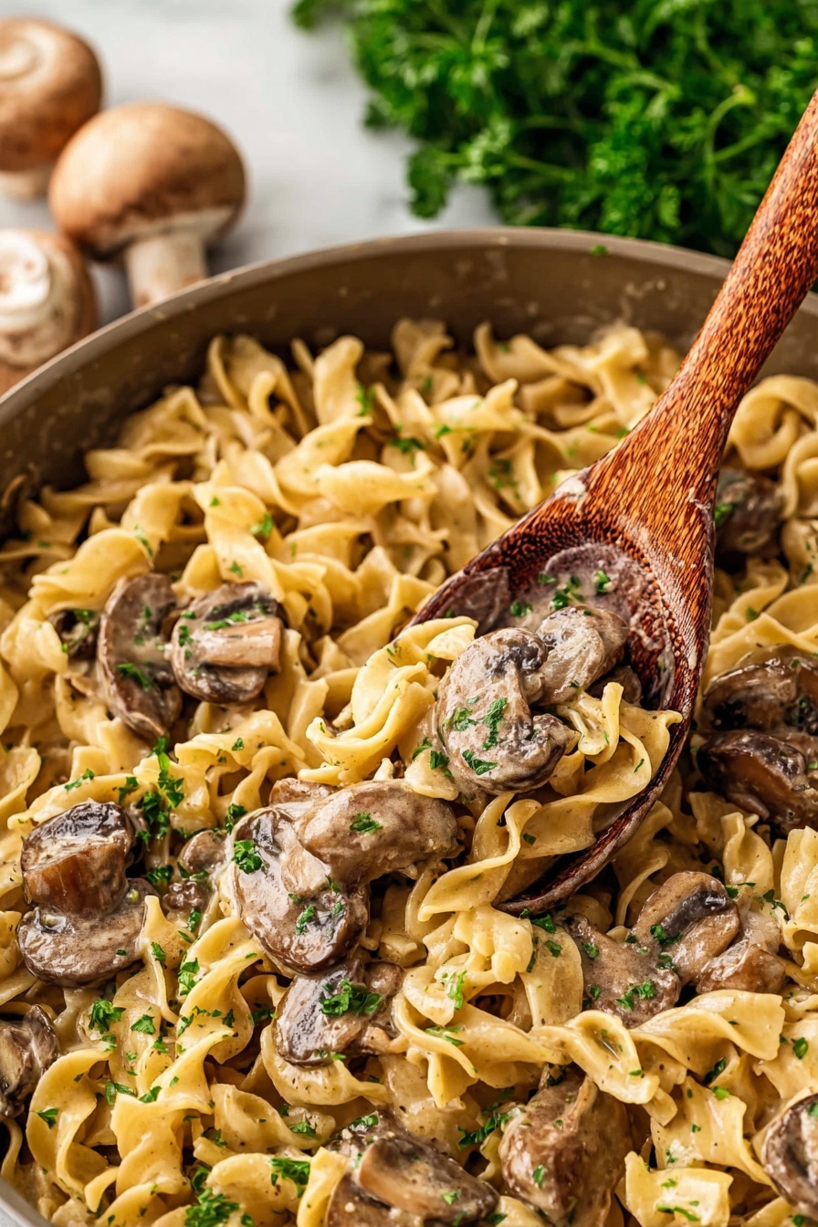 Old Fashioned Beef Stroganoff, beef stroganoff recipe, classic beef stroganoff, creamy beef stroganoff, easy beef stroganoff - The image shows a close-up of a pan filled with a creamy noodle and mushroom dish. There are egg noodles that are light yellow, soft, and curly, spread all over the pan as the first layer. On top of the noodles, there are sliced mushrooms with a brown color and slightly shiny surface from the sauce. Mixed with the mushrooms and noodles, there are small bits of green parsley scattered for color. A wooden spoon is scooping some of the dish from the pan. The background has some blurred green parsley and whole mushrooms placed on a white marbled surface. photo taken with an iphone --ar 2:3 --v 7