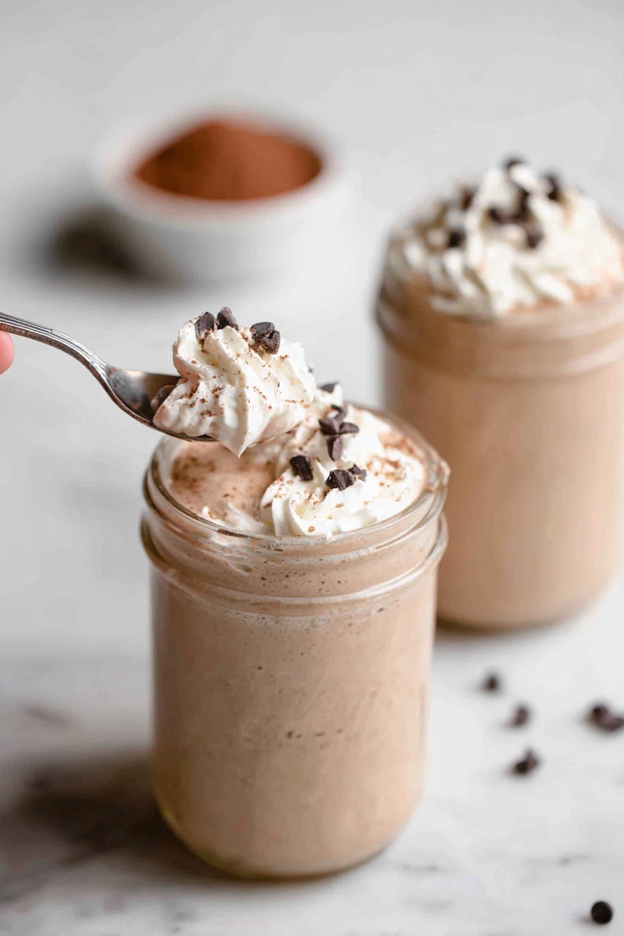 Vegan Protein Shake, plant-based protein smoothie, healthy vegan breakfast, dairy-free protein shake, quick vegan shake - Two glass jars sit on a white marbled surface, each filled with a creamy light brown milkshake. The milkshakes have a thick texture and are topped with a swirl of white whipped cream. Small, dark chocolate chips are sprinkled over the whipped cream. In the foreground, a silver spoon held by a woman's hand scoops a portion of whipped cream and chocolate chips from the first jar. The background jar is out of focus but shows the same whipped cream and chocolate chip topping. There is a small bowl filled with cocoa powder blurred in the background. Photo taken with an iphone --ar 2:3 --v 7