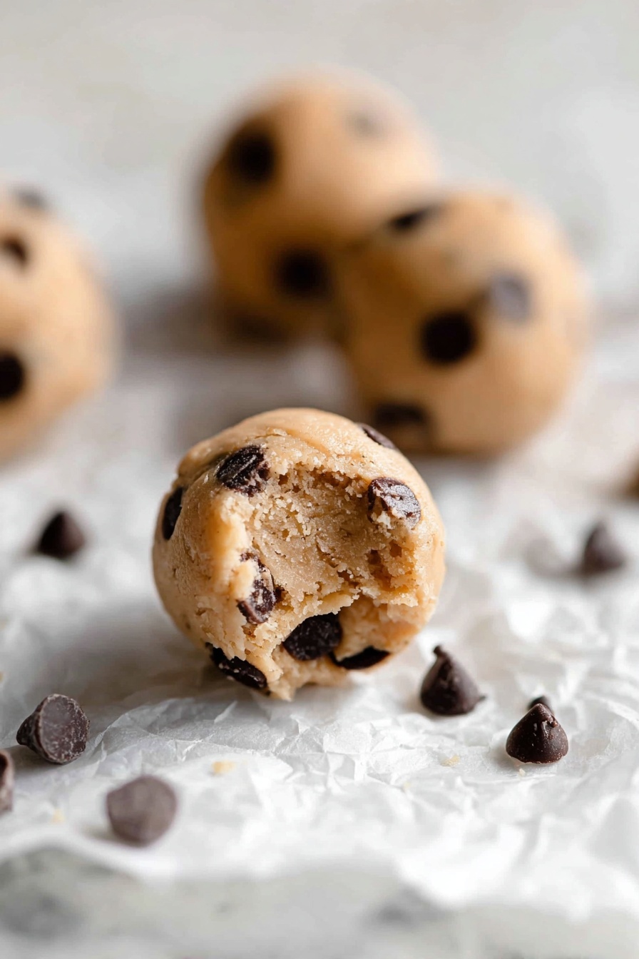 Protein Cookie Dough Bites, healthy cookie dough bites, no-bake protein snacks, high-protein dessert, quick healthy treats - The image shows small round cookie dough balls with a light tan color, filled with many dark brown chocolate chips scattered inside and on the surface. The balls rest on a crumpled white paper on a white marbled background. One dough ball in the front has a small bite taken out of it, showing a soft, slightly crumbly texture inside, while other dough balls and some loose chocolate chips are blurred in the background. The lighting is soft and natural, highlighting the texture of the dough and chocolate chips, photo taken with an iphone --ar 2:3 --v 7