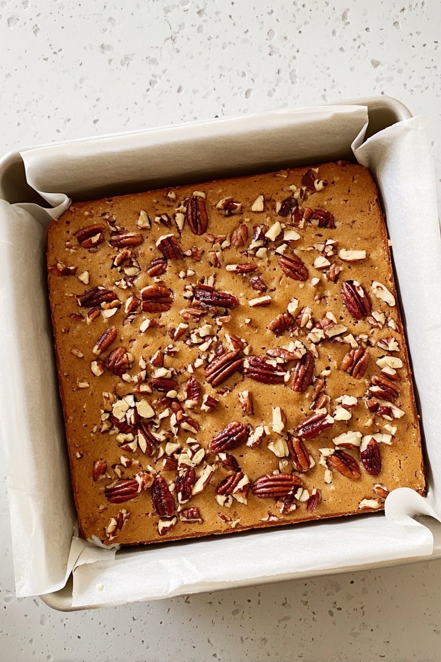 Maple Pecan Blondies, maple blondie recipe, nutty blondie bars, caramel pecan blondies, buttery dessert bars - A square baked dessert with a light brown top covered in scattered pieces of pecans. It is in a white baking pan lined with parchment paper that folds over the edges. The baked item looks soft and slightly puffy, with a smooth but slightly textured surface from the nuts. The pan sits on a white marbled surface. photo taken with an iphone --ar 2:3 --v 7