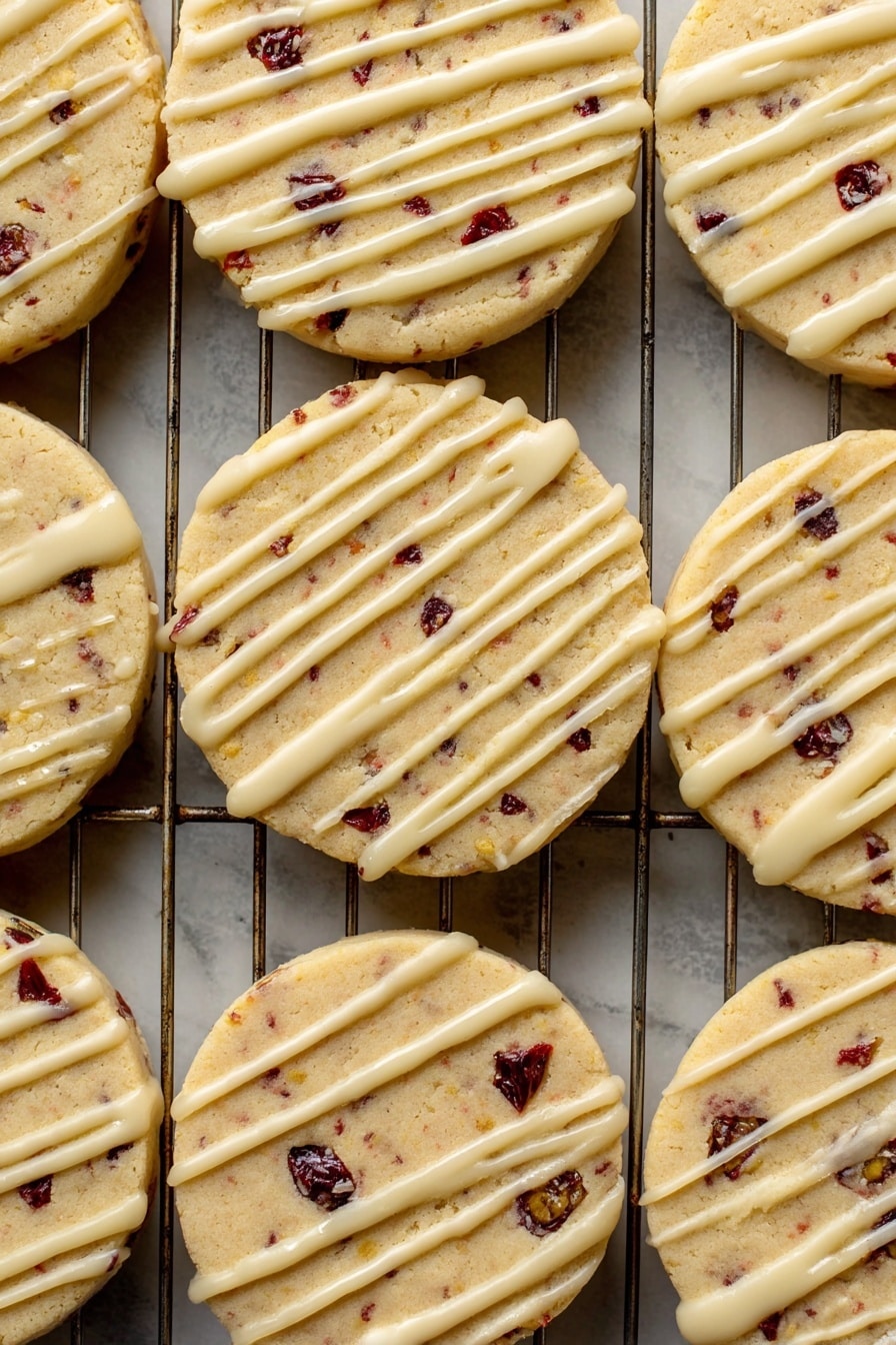 Orange Cranberry Shortbread Cookies, orange cranberry cookies, zesty shortbread recipes, vegan shortbread cookies, holiday cookie recipes - A close-up view of a batch of round cookies laid out on a metal cooling rack over a white marbled surface. Each cookie has one visible layer, light golden in color with small dark red and black fruit bits inside, giving a spotted look. On top of each cookie is a thin, even drizzle of pale cream-colored icing, applied unevenly in long lines across the surface. The texture of the cookies looks soft but firm, with some slight cracks near the edges. The metal wires of the rack create a grid pattern underneath the cookies. Photo taken with an iphone --ar 2:3 --v 7