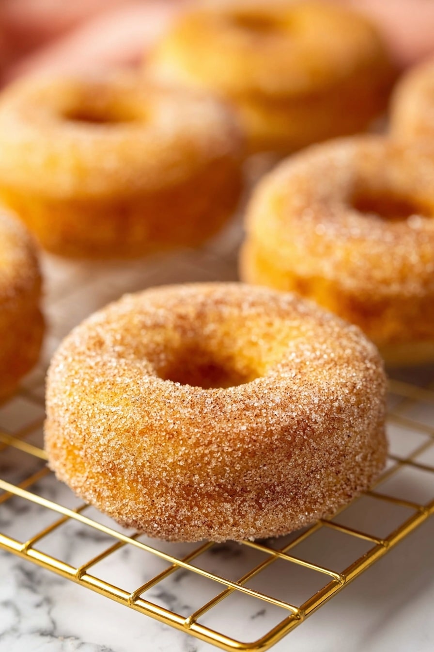Vegan Apple Cider Donuts, vegan fall donuts, vegan apple cider treats, easy vegan donut recipe, cozy fall dessert - The image shows several round donuts with a hole in the middle, each covered with a layer of fine sugar and cinnamon on the outside. The donuts have a golden-brown color and a soft texture. They are placed on a gold wire cooling rack, sitting on a white marbled surface. The focus is on one donut in the front, with other donuts slightly blurred in the background, giving a warm and fresh look. photo taken with an iphone --ar 2:3 --v 7