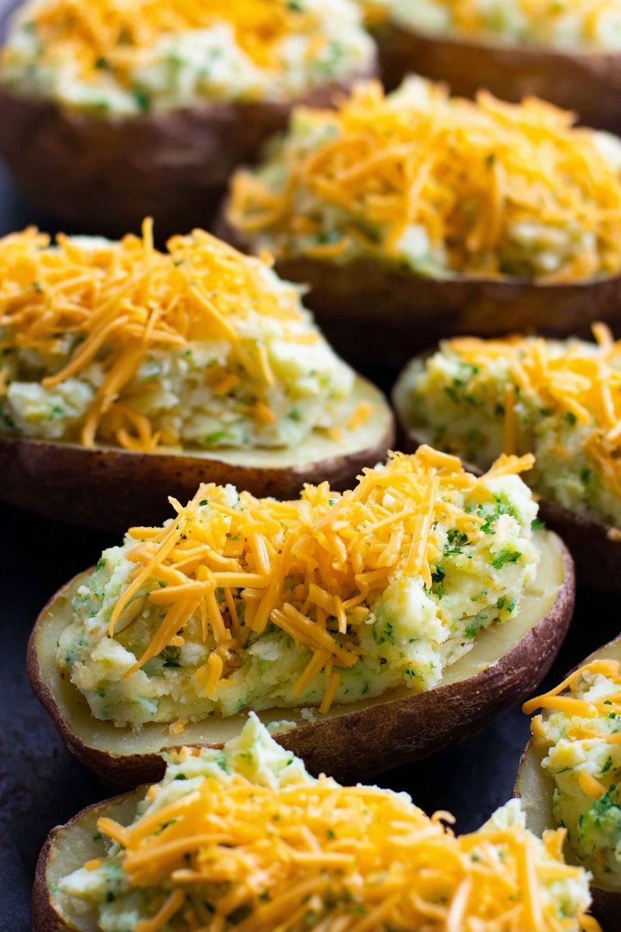 Vegan Cheesy Broccoli Twice Baked Potatoes, vegan stuffed baked potatoes, plant-based cheesy potato recipes, dairy-free broccoli potatoes, vegan comfort food ideas - The image shows several potato halves with the inside scooped out and filled with a creamy mixture that looks light yellow with green bits throughout, suggesting herbs or vegetables mixed in. Each stuffed potato is topped with a generous layer of shredded orange cheddar cheese. The potatoes are arranged closely together on a dark surface which contrasts with the light filling and bright cheese. The texture of the filling appears smooth but slightly chunky, and the potato skins are a dark brown color. The close-up shot highlights the layers clearly with the cheese on top, the creamy filling in the middle, and the potato skin at the bottom. Photo taken with an iphone --ar 2:3 --v 7