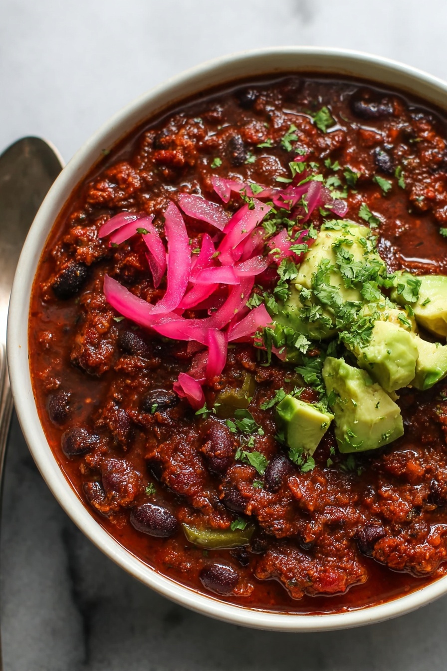 Lentil Chili, vegetarian chili recipe, vegan chili, healthy lentil stew, easy chili recipe - A close-up view of a bowl of thick chili, showing three main layers: a base layer of rich, dark red-brown chili with black and red beans and small pieces of green bell pepper spread throughout, topped on one side with bright pink pickled onion slices, and on the same side, fresh green avocado chunks sprinkled with finely chopped cilantro. The bowl is white and a spoon is partially visible on the left side, all placed on a white marbled surface. Photo taken with an iphone --ar 2:3 --v 7