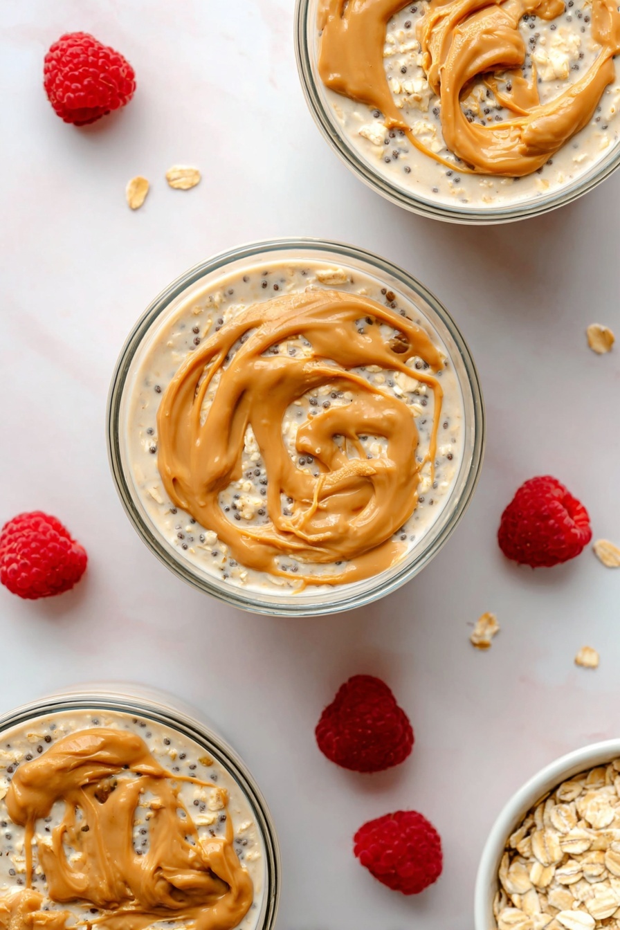 Peanut Butter Overnight Oats, healthy breakfast ideas, easy overnight oats, quick morning meal, creamy peanut butter oats - The image shows three round clear glass bowls filled with layered overnight oats on a white marbled surface. Each bowl has a bottom layer of creamy light beige oats mixed with small black chia seeds, covered by a thin layer of milky liquid. The top layer is swirled with dollops of light brown peanut butter, creating smooth, curved textures on the surface. Around the bowls, scattered fresh bright red raspberries add a pop of color, along with a small white bowl filled with rolled oats placed in the bottom right corner. The photo taken with an iphone --ar 2:3 --v 7