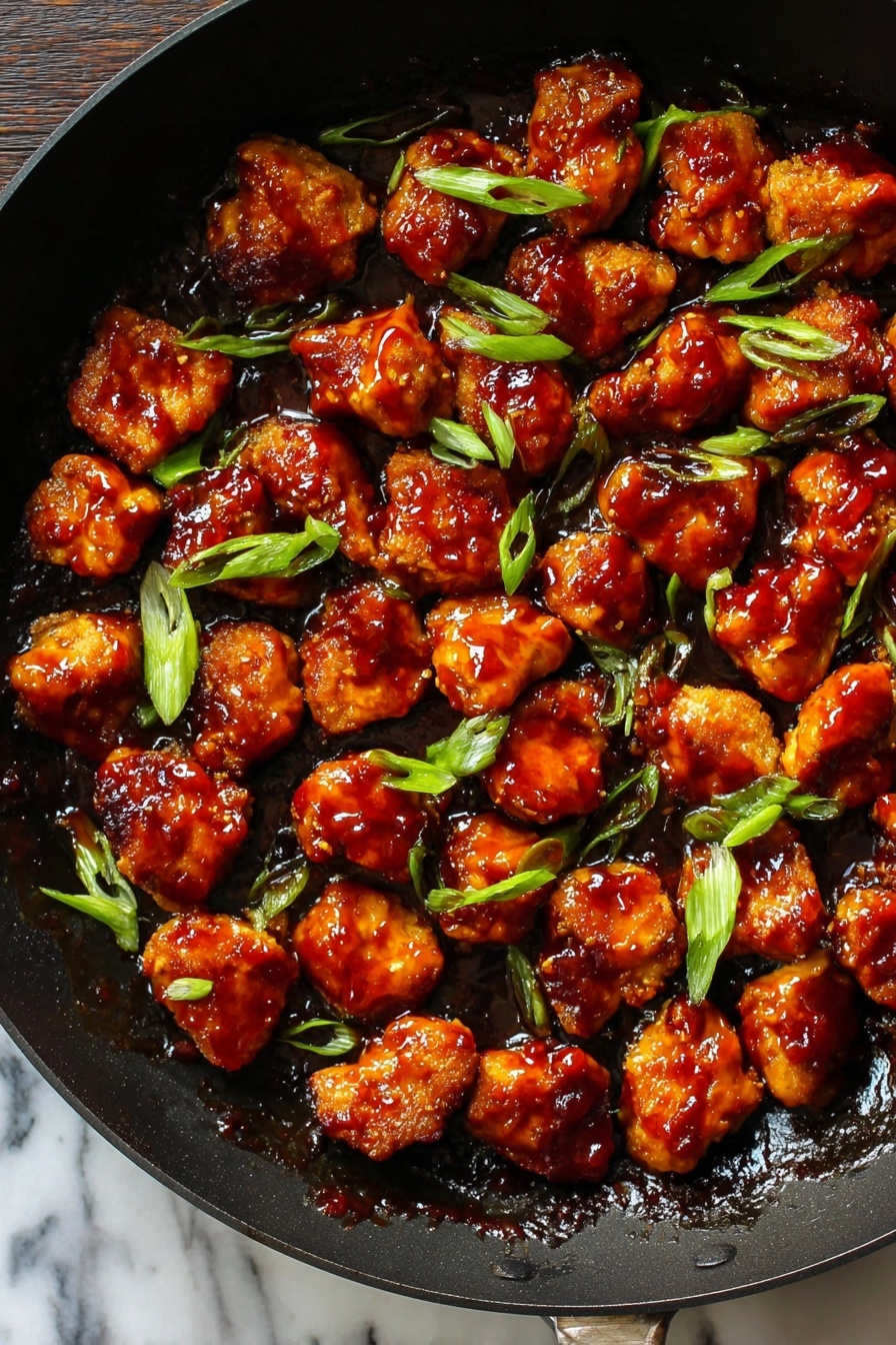 Honey Ginger Seitan with Crispy Pan-Fried Coating, vegan seitan recipes, crispy vegan protein, homemade seitan with ginger, savory vegan dinner ideas - This image shows a black pan filled with many small pieces of golden-brown chicken covered in thick, shiny reddish-brown sauce. The pieces are spread out evenly across the pan, with some bright green slices of scallion sprinkled on top. The sauce looks sticky and glossy, covering all the chicken pieces in a rich layer. The pan's dark color contrasts with the vibrant colors of the chicken and scallions. The background surface is a white marbled texture. photo taken with an iphone --ar 2:3 --v 7