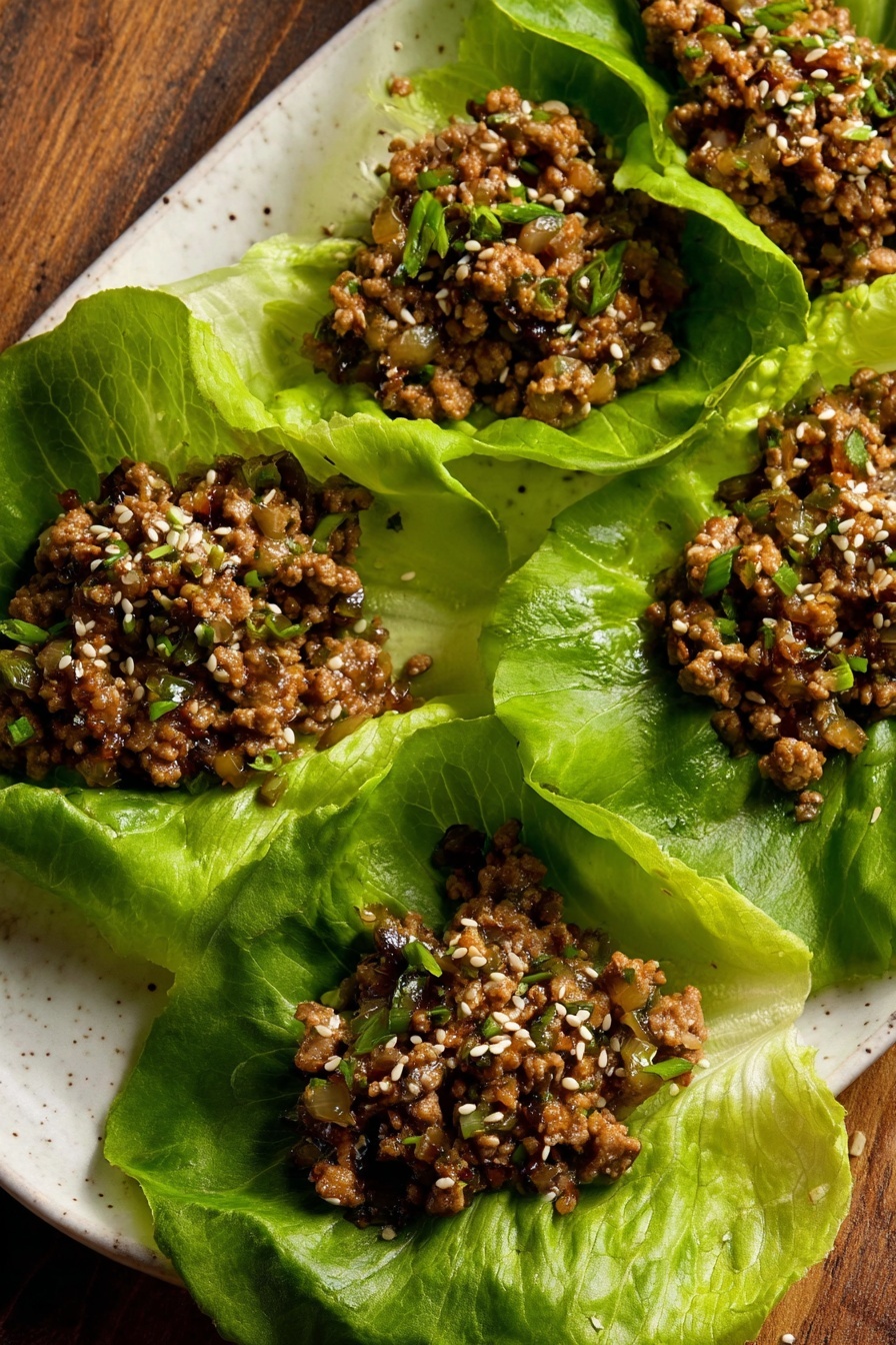 Tofu Lettuce Wraps, healthy tofu lettuce wraps, vegan lettuce wraps, easy tofu wraps, crispy tofu recipes - The image shows five large green lettuce leaves arranged on a white plate with a wood surface in the background. Each leaf holds a serving of cooked ground meat mixed with small diced vegetables, with a brown color and a slightly moist texture. The meat mixture is sprinkled with white sesame seeds and small bits of green herbs. The lettuce leaves are fresh with visible veins and crinkled edges, creating a bright green contrast to the darker meat filling. Photo taken with an iphone --ar 2:3 --v 7