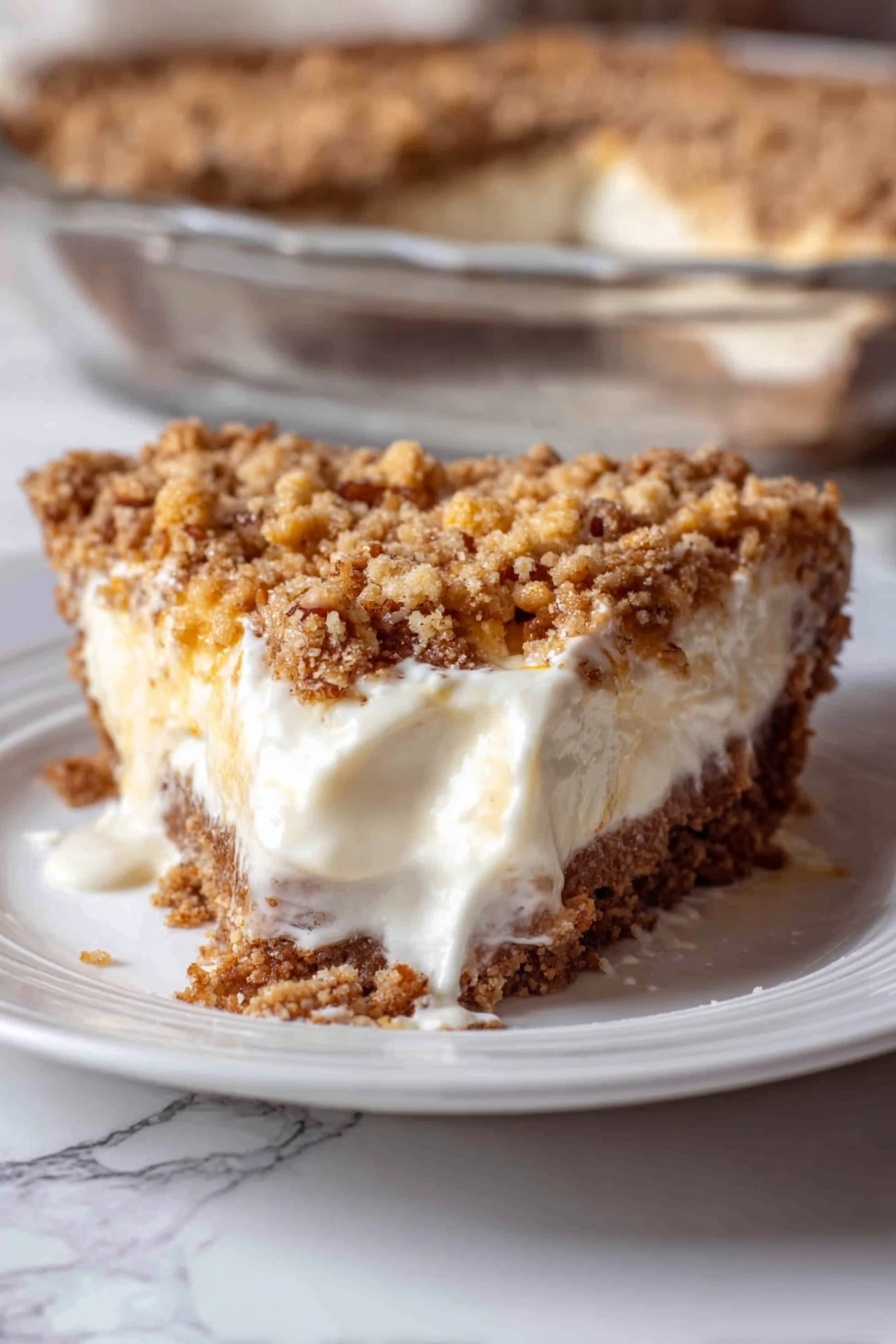 Angel Pecan Pie, light pecan pie, fluffy pecan pie, crunchy cracker crust pecan pie, festive pecan pie dessert - A close-up of a slice of pie on a white plate with a white marbled surface underneath. The pie has three visible layers: a crumbly brown crust at the bottom, a thick creamy white filling in the middle that is slightly melting and spreading on the plate, and a chunky golden brown crumb topping with some small nut bits on top. In the background, part of a clear glass dish shows the rest of the pie with the same three layers. The photo taken with an iphone --ar 2:3 --v 7