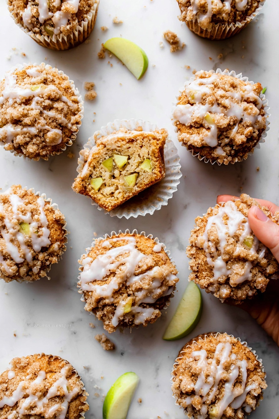 Apple Cinnamon Crumb Muffins, apple cinnamon muffins, cozy breakfast recipes, apple muffin recipe, cinnamon crumb muffins - The image shows nine apple crumb muffins arranged on a white marbled surface. Each muffin has three clear layers: a bottom baked golden-brown cake layer, a middle layer with visible light green apple chunks inside the muffin, and a top crumbly light brown streusel topping with a slight sprinkle of white glaze drizzled unevenly over it. One muffin is open, showing the inside texture and apple pieces, sitting in a white paper liner with a woman's hand about to pick it up. Small crumbs and a few green apple slices are scattered around, adding detail to the scene. Photo taken with an iphone --ar 2:3 --v 7