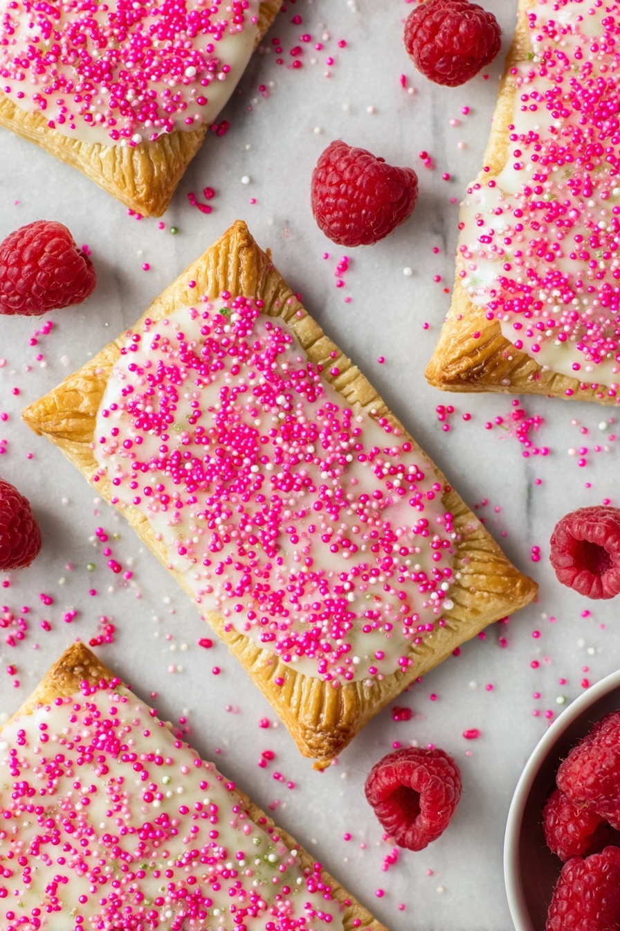 Raspberry Pop Tarts, homemade raspberry breakfast pastries, flaky fruit-filled tarts, easy homemade pop tarts, raspberry pastry recipes - Several rectangular toasted pastries spread over a white marbled surface, each with a golden-brown crust stamped with fork marks along the edges; the top layer is coated in a slightly shiny white icing sprinkled generously with bright pink sugar crystals, creating a textured look. Scattered around the pastries are fresh, plump red raspberries adding a pop of natural color. In the bottom right corner, a small portion of a white bowl is visible, holding more raspberries. photo taken with an iphone --ar 2:3 --v 7