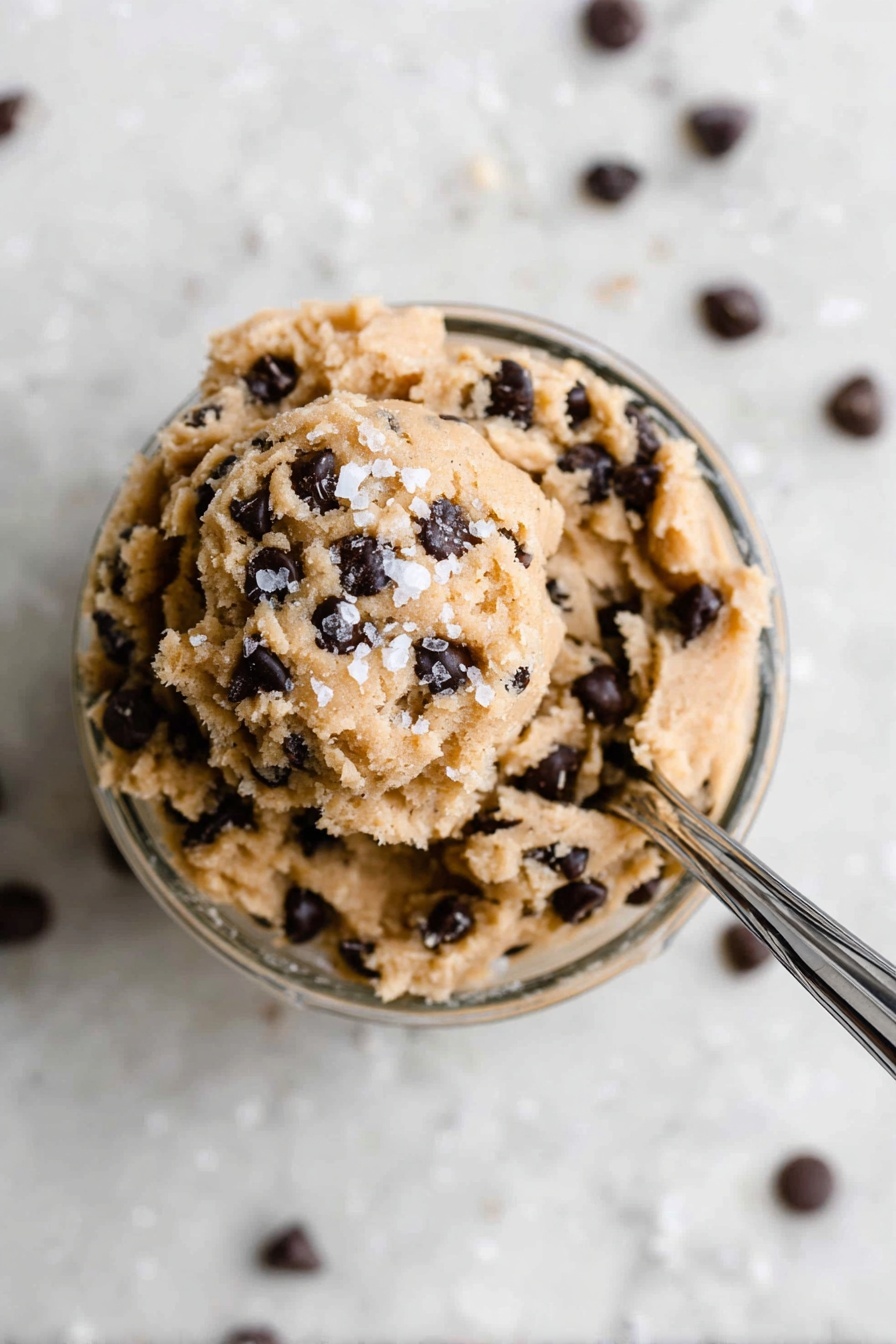Protein Cookie Dough Bites, healthy cookie dough bites, no-bake protein snacks, high-protein dessert, quick healthy treats - In a small clear glass bowl filled with one layer of light brown, soft cookie dough mixed with many small dark chocolate chips all over, the dough is slightly scooped at the top with a few coarse white salt flakes sprinkled on it. A shiny silver spoon is stuck into the dough from the right side, angled toward the top right corner, resting inside the bowl. The bowl sits on a surface with a white marbled texture, and some chocolate chips are scattered around it. The photo is taken with an iphone --ar 2:3 --v 7