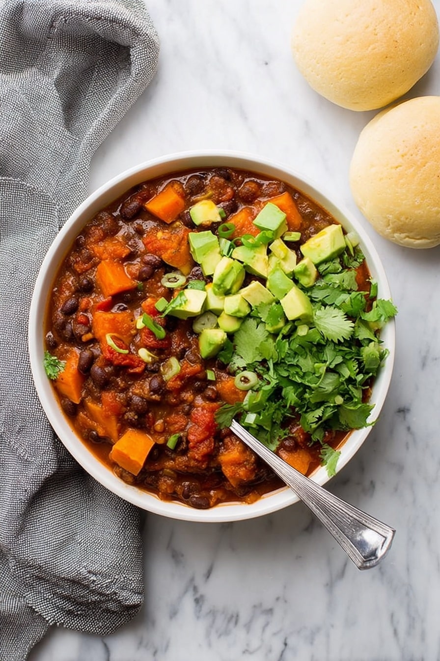 Sweet Potato Black Bean Chili, healthy plant-based chili, hearty vegan chili, easy vegetarian chili, cozy winter chili - A white bowl filled with three main layers: the base is a thick stew of dark brown beans mixed with deep orange chunks of sweet potato and red tomato pieces in a reddish-brown sauce, topped on one side with bright green chopped avocado cubes and sprinkled with small rings of green onion, and on the other side with fresh green cilantro leaves. A silver spoon rests inside the bowl at the right side. The bowl sits on a white marbled surface next to two light beige round bread rolls and a loosely folded gray cloth. Photo taken with an iphone --ar 2:3 --v 7