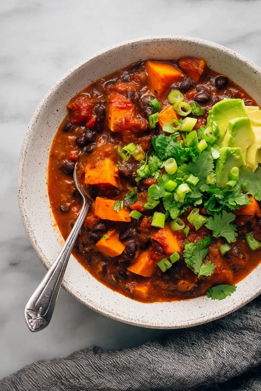 Sweet Potato Black Bean Chili, healthy plant-based chili, hearty vegan chili, easy vegetarian chili, cozy winter chili - The image shows a white speckled bowl filled with a thick stew made of dark black beans and bright orange chunks of sweet potato in a rich red tomato sauce. The stew is garnished with vibrant fresh green cilantro leaves and sliced green onions, while creamy green avocado pieces sit on one side near the bowl’s edge. A silver spoon is placed in the stew with its handle resting on the edge of the bowl. The bowl is set on a white marbled surface with a gray cloth draped nearby. photo taken with an iphone --ar 2:3 --v 7
