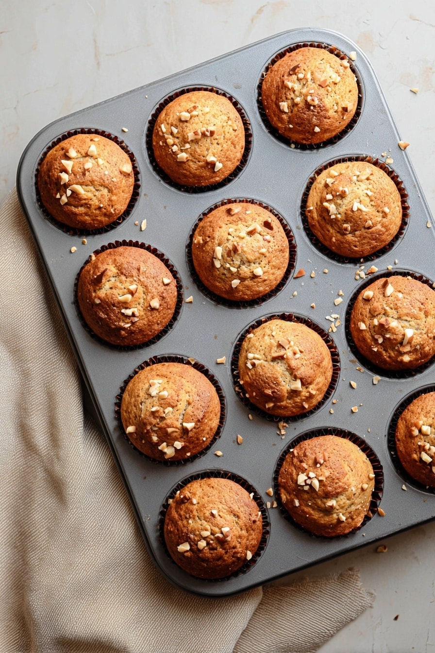 Quick & Easy Vegan Banana Muffins, vegan banana muffins, healthy vegan muffins, fluffy banana muffins, quick vegan baking - A grey muffin tray holds twelve freshly baked muffins, each with a golden-brown top that is slightly rounded and textured with small cracks. Light tan chopped nuts are scattered on the surface of each muffin, adding a crunchy detail. The muffins sit in dark brown paper liners that are barely visible around the edges. The tray rests on a white marbled texture surface with a soft beige cloth partially visible on the left side. The light gently highlights the warm tones and textures of the muffins, making them look soft and fresh. photo taken with an iphone --ar 2:3 --v 7