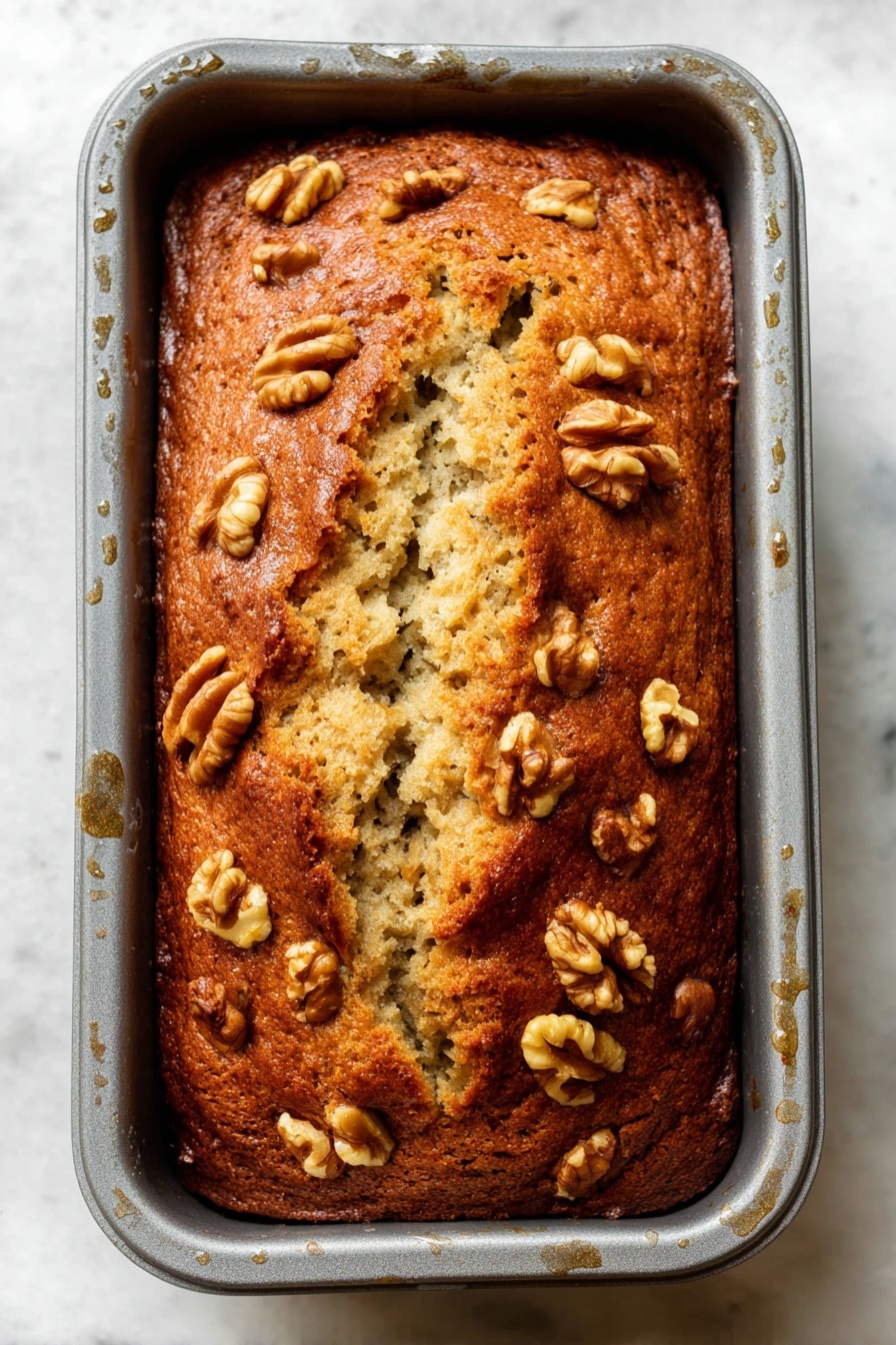 Best Vegan Banana Bread, vegan banana bread recipe, plant-based banana bread, moist banana bread, vegan baking recipes - A loaf of golden brown banana bread with a slightly cracked top sits inside a gray baking pan, showing the soft, light beige interior through the cracks. The surface is sprinkled with medium-sized walnut halves that are toasted to a light brown, scattered evenly on top and around the edges. The loaf has a dense, moist texture with visible bits of banana inside, and the baking pan has some visible moisture droplets on its sides. The background is a white marbled texture. Photo taken with an iphone --ar 2:3 --v 7