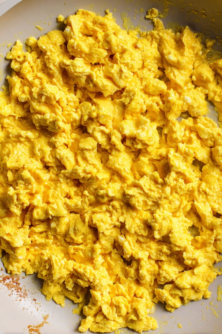 Tofu Scramble, vegan breakfast, healthy plant-based recipes, easy tofu scramble, flavorful vegan breakfast - The image shows a close-up view inside a white pan filled with scrambled eggs. The cooked eggs have a soft, slightly chunky texture with a bright yellow color spread evenly across the pan. The edge of the pan reveals some lightly browned bits where the eggs touched the surface. The background is a white marbled texture. photo taken with an iphone --ar 2:3 --v 7