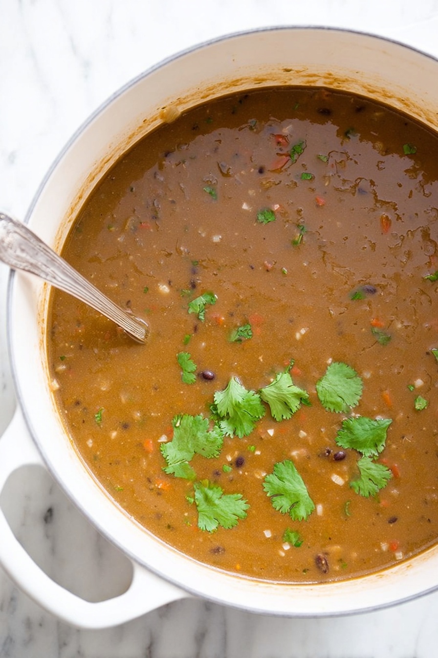 Vegan Black Bean Soup, vegan black bean soup recipe, hearty vegan soup, easy black bean soup, plant-based black bean stew - A large white pot filled with thick brown soup that has a slightly grainy texture, small visible pieces of vegetables, and beans mixed throughout. Several fresh green cilantro leaves float on top, adding a touch of bright green color. A silver spoon is partially dipped into the soup on the left side of the pot. The pot is placed on a white marbled surface. Photo taken with an iphone --ar 2:3 --v 7
