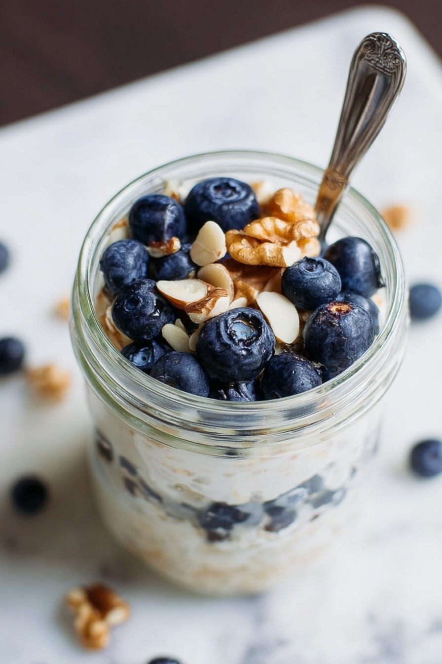Simple Vegan Overnight Oats, vegan overnight oats, healthy breakfast ideas, quick vegan breakfast, customizable overnight oats - A clear glass jar filled with three layers: the bottom layer is fresh blueberries, the middle layer is creamy white porridge or yogurt with a smooth texture, and the top layer has more blueberries and a few light brown walnut pieces scattered on top. A silver spoon is placed in the jar, partly inside the food. The surface around the jar shows a white marbled texture with some scattered white nut pieces. Photo taken with an iphone --ar 2:3 --v 7