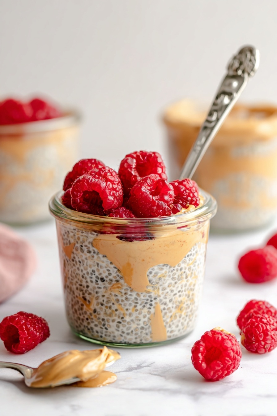 Peanut Butter Overnight Oats, healthy breakfast ideas, easy overnight oats, quick morning meal, creamy peanut butter oats - A clear glass jar filled with three visible layers: the bottom layer is light beige chia seed pudding with black chia seeds mixed in, the middle layer shows swirls of creamy light brown peanut butter, and the top layer is a bright red cluster of fresh raspberries. A silver spoon with an ornate handle is placed inside the jar, leaning against the side with some pudding on it. Around the jar, scattered fresh raspberries add more red spots on a white marbled surface. In the background, more jars with similar contents and a silver spoon with a light brown spread are softly blurred. Photo taken with an iphone --ar 2:3 --v 7