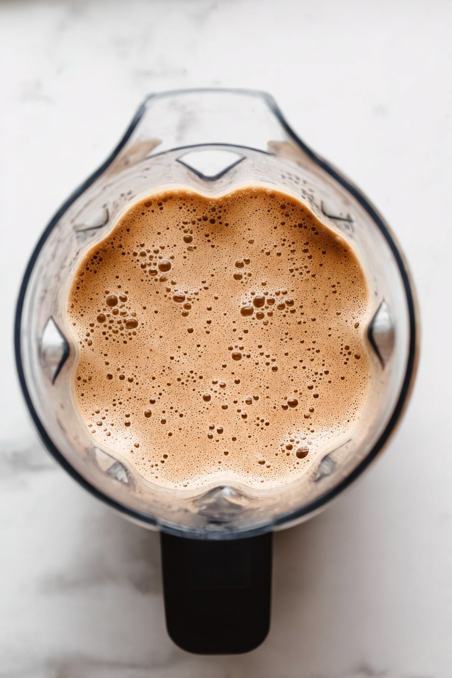 Vegan Protein Shake, plant-based protein smoothie, healthy vegan breakfast, dairy-free protein shake, quick vegan shake - The image shows a clear blender from above filled with a light brown, frothy mixture with many small bubbles on its surface. The blender has a black handle, and the background is a white marbled surface. photo taken with an iphone --ar 2:3 --v 7