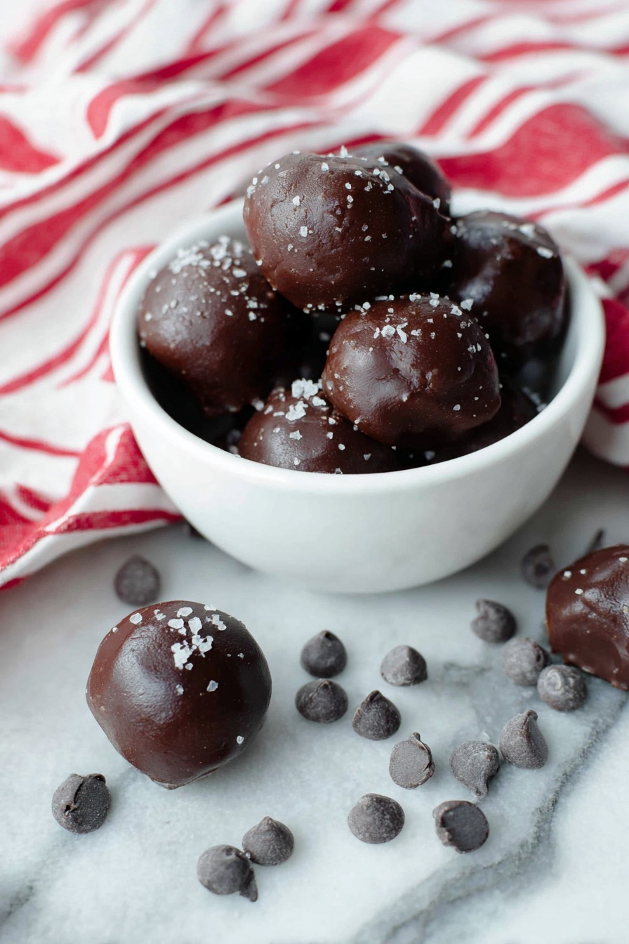 Cocoa Date Truffles, healthy chocolate truffles, natural sweetened treats, vegan dessert ideas, easy chocolate truffles - A white bowl filled with several dark brown chocolate balls that have a smooth, shiny texture and are sprinkled lightly with coarse salt flakes on top. Around the bowl, more chocolate balls sit on a white marbled surface, some resting near small clusters of dark chocolate chips. A white cloth with bold red stripes is casually draped to the back and right side of the bowl, adding a bright contrast. The scene is bright and clean, showing the rich dark color and glossy texture of the chocolates clearly. Photo taken with an iphone --ar 2:3 --v 7