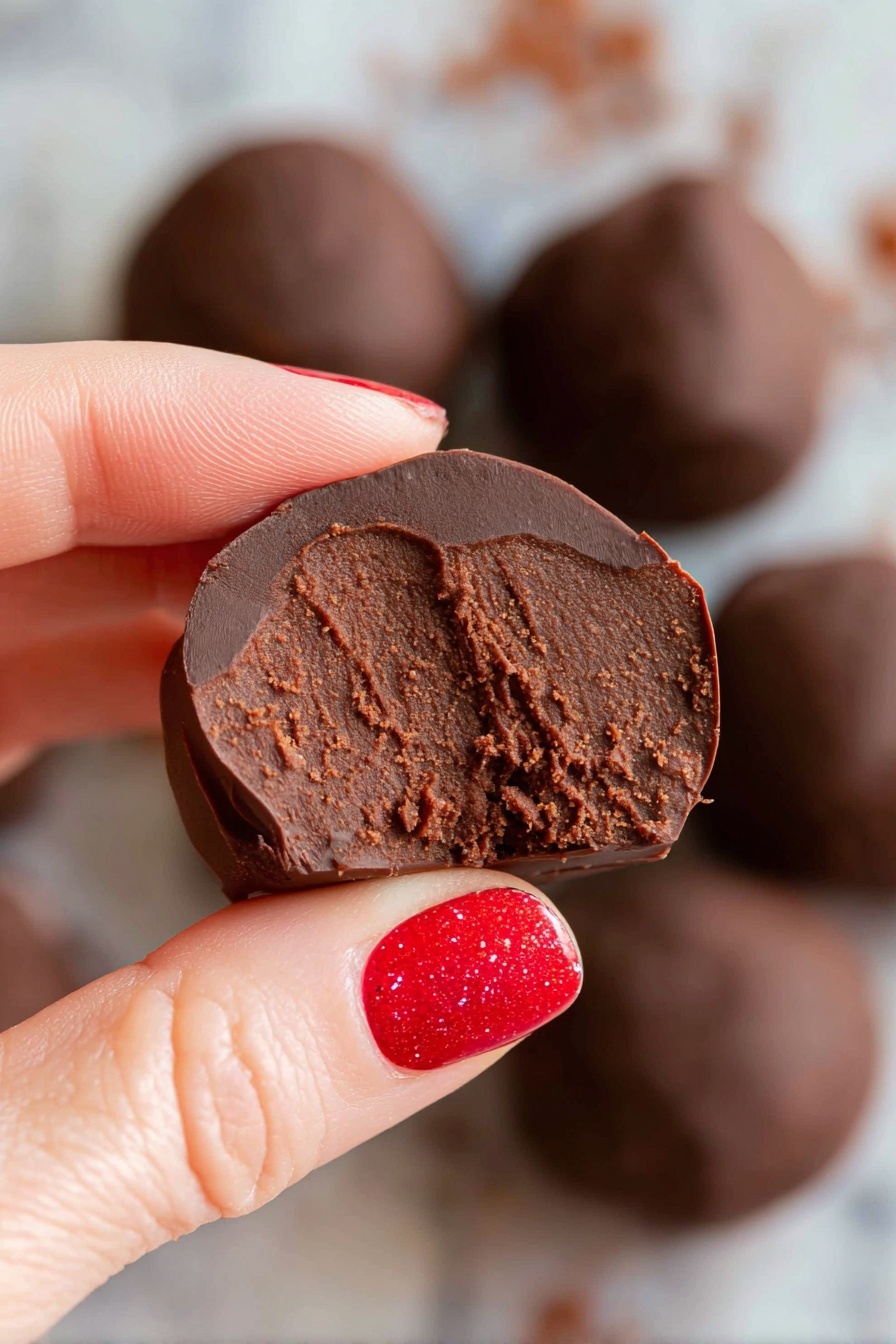 Healthy Chocolate Bean Snack Balls, chocolate bean snack balls, wholesome chocolate protein bites, healthy chocolate treats, quick healthy snack ideas - A round white bowl filled with several smooth, shiny dark chocolate balls stacked together. Two of the chocolate balls are cut in half and placed on top, revealing a dense, rich, and slightly grainy brown chocolate filling inside. The bowl sits on a white marbled surface, giving a clean and bright look. The chocolate balls have a glossy outer layer with some small wrinkles, showing a contrast between the shiny dark coating and textured inside. Photo taken with an iphone --ar 2:3 --v 7