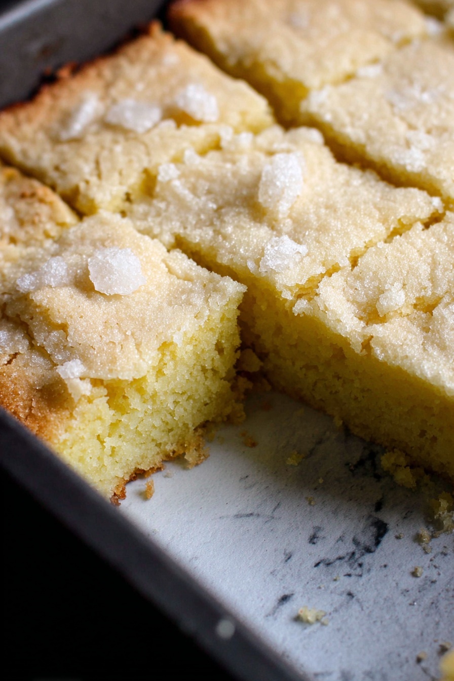 Creamy Butter Cake, buttery cake recipe, moist vanilla cake, classic cake with butter, fluffy butter cake - A single-layer light yellow cake is shown inside a dark baking pan with a white marbled texture surface underneath. The top layer has a rough crumbly texture with sugar crystals scattered on top, and it is cut into square pieces with one piece missing, revealing a soft, dense inside that is smooth and slightly moist. The cake edges are slightly browned and cracked, contrasting with the pale yellow center. Photo taken with an iphone --ar 2:3 --v 7