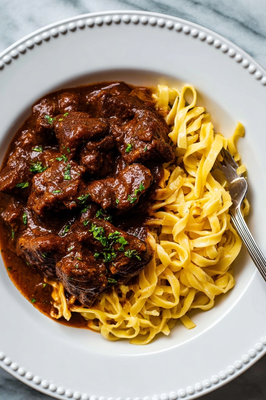 German Beef Goulash, hearty beef goulash recipe, authentic German goulash, easy beef goulash, comfort food German dish - A white plate with a raised dotted edge holds a dish of thick yellow noodles on the bottom right, with a silver fork resting on them. On top and mostly to the left of the noodles, there are large dark brown chunks of meat in rich brown sauce. The sauce is thick and covers most of the meat, with small pieces of green herbs sprinkled over the meat and noodles. The plate is placed on a white marbled surface. photo taken with an iphone --ar 2:3 --v 7