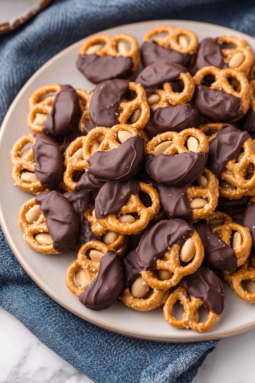 No-Bake Peanut Butter Pretzel Bites, easy no-bake snacks, salty sweet treats, quick dessert ideas, chocolate peanut butter bites - A white plate filled with many small pretzels, each topped with a single peanut and half dipped in smooth dark chocolate, giving a mix of shiny black and golden tan colors. The pretzels have a crunchy texture with visible salt grains on the surface, while the peanuts underneath add a light brown soft contrast. The plate sits on a white marbled texture with a blue cloth nearby. photo taken with an iphone --ar 2:3 --v 7