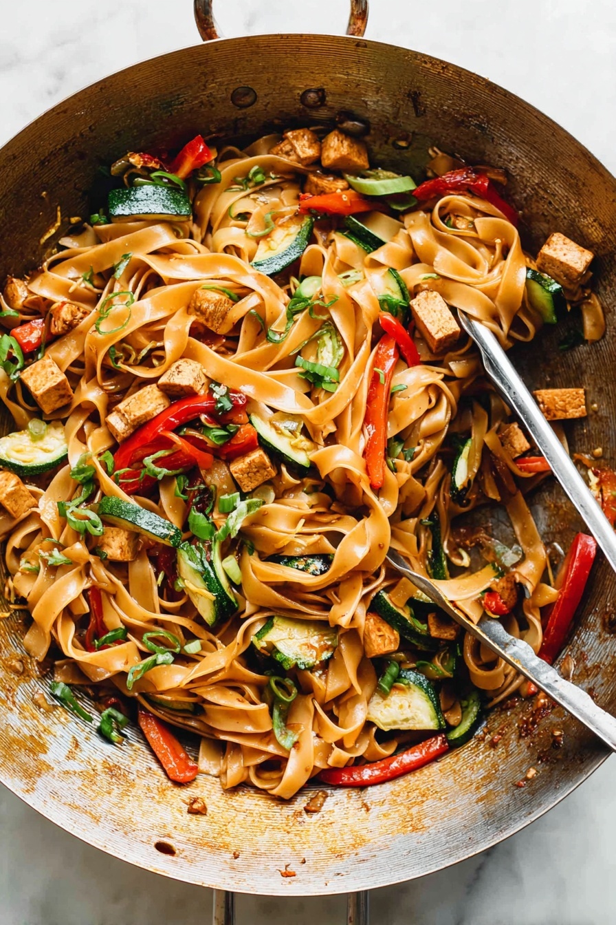 Drunken Noodles, Thai stir-fry recipe, spicy noodle dish, quick dinner recipe, flavorful Asian noodles - The image shows a metal wok filled with a colorful stir-fry dish. The base layer consists of wide, flat noodles that are light brown and glossy. Mixed through the noodles are pieces of lightly browned tofu cubes, thin strips of green bell peppers, slices of red bell peppers, and small chunks of zucchini with their green skin visible. Scattered over the top are thinly sliced green onions and bits of chopped herbs adding dots of bright green. The noodles and vegetables appear shiny from a light sauce coating them evenly, and the wok has a warm, well-used appearance with some browning on the sides. A pair of metal tongs rests inside the wok, ready to serve. The setting has a clean, white marbled texture surface. photo taken with an iphone --ar 2:3 --v 7