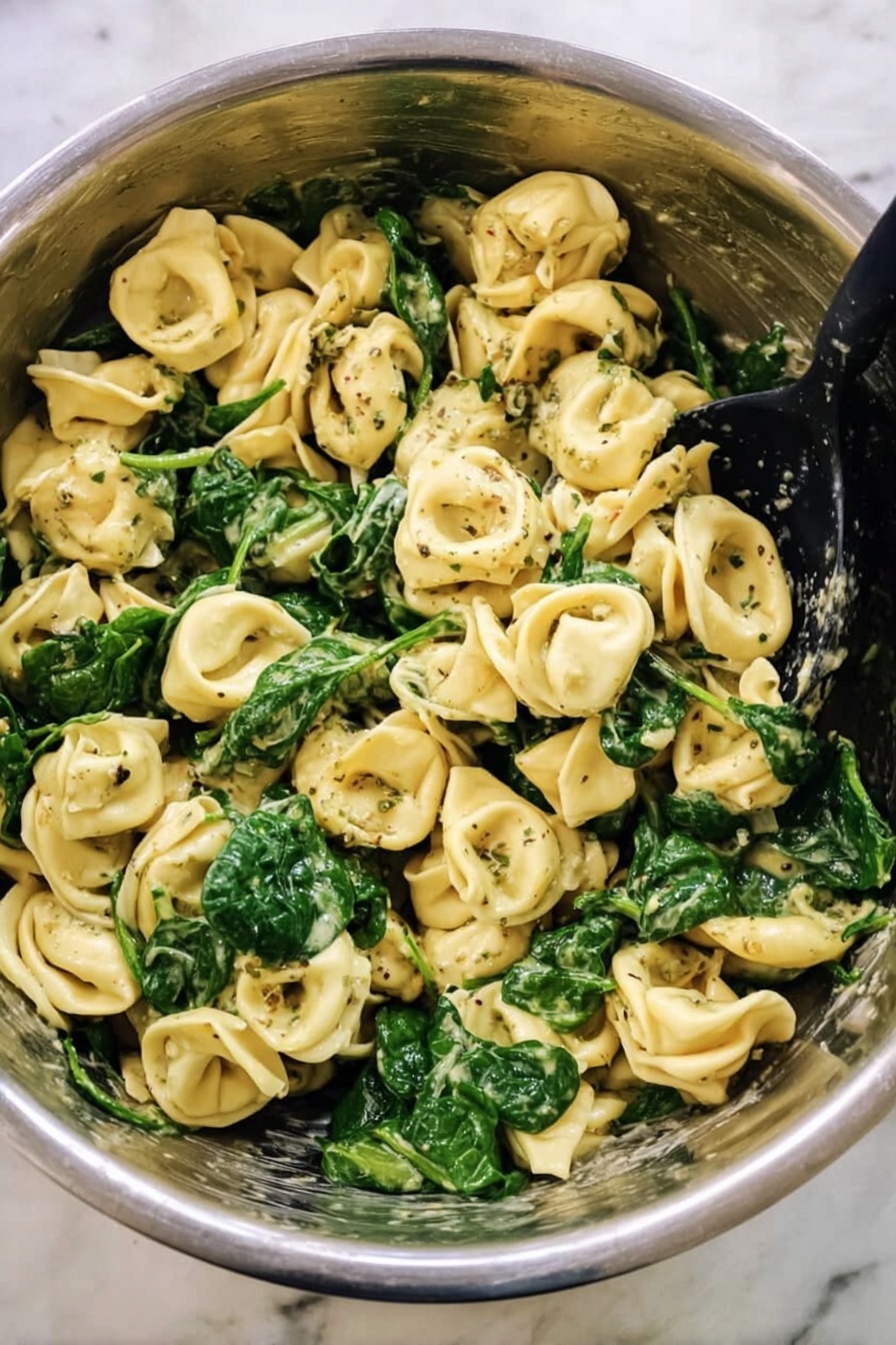 Cheese Tortellini with Pesto and Spinach, quick pasta recipes, easy vegetarian dinner, healthy tortellini ideas, Italian pasta with pesto - This image shows a big metal bowl filled with cooked tortellini pasta mixed with bright green spinach leaves. The pasta is light yellow with a soft texture, and some pieces have dark green herb specks on them. The spinach looks fresh and slightly wilted, spread evenly throughout the pasta. There is a black spoon resting inside the bowl, ready to serve. The bowl sits on a white marbled surface, giving a clean and simple background. Photo taken with an iphone --ar 2:3 --v 7