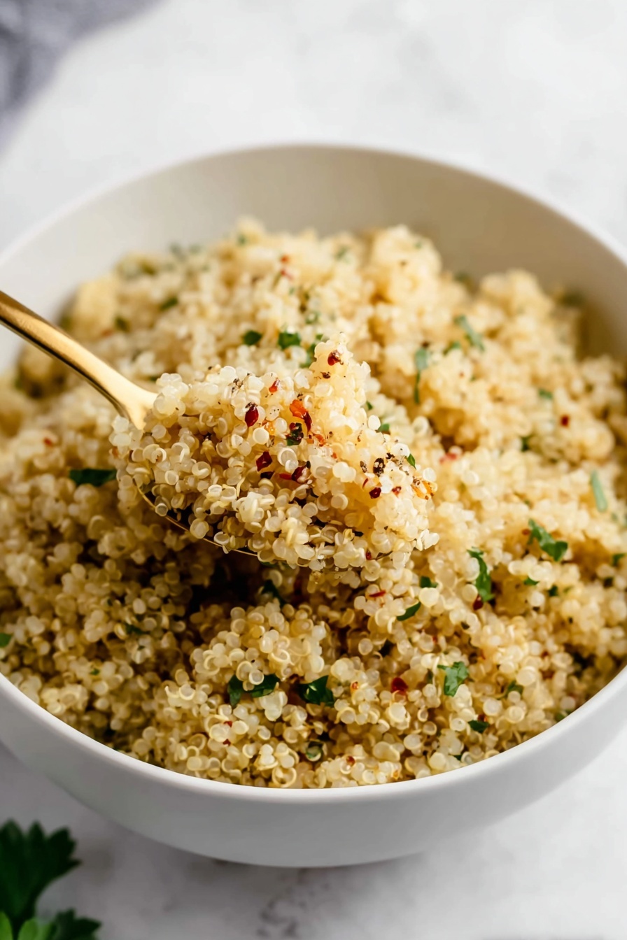 Garlic Butter Quinoa, garlic butter quinoa side dish, easy garlic quinoa recipe, savory quinoa recipes, quick vegetarian quinoa - A close-up of a white bowl full of cooked quinoa, which is light beige and fluffy. The quinoa grains are mixed with small green herb pieces scattered evenly throughout and seasoning specks of black and red on top. A gold spoon scoops up some quinoa from the bowl, showing the grain’s soft, slightly textured surface. The bowl sits on a white marbled texture surface with some green leaves blurred in the background. photo taken with an iphone --ar 2:3 --v 7