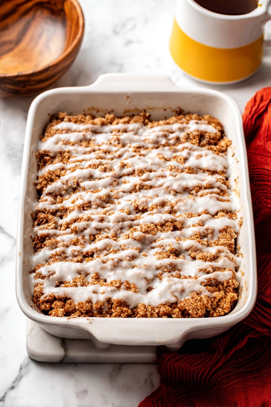 Pumpkin Crumb Coffee Cake, pumpkin coffee cake with crumb topping, fall pumpkin breakfast cake, moist pumpkin coffee cake recipe, easy pumpkin coffee cake - The image shows a white rectangular baking dish filled with a crumbly brown dessert layer, topped with a drizzle of white icing spread unevenly across the surface. The crumb layer has a soft, rough texture with patches of darker and lighter brown. The dish is placed on a white marbled surface, with part of a wooden bowl visible on the left and a yellow and white mug on the right. A red cloth lies partially beneath the baking dish at the bottom right corner. Photo taken with an iphone --ar 2:3 --v 7