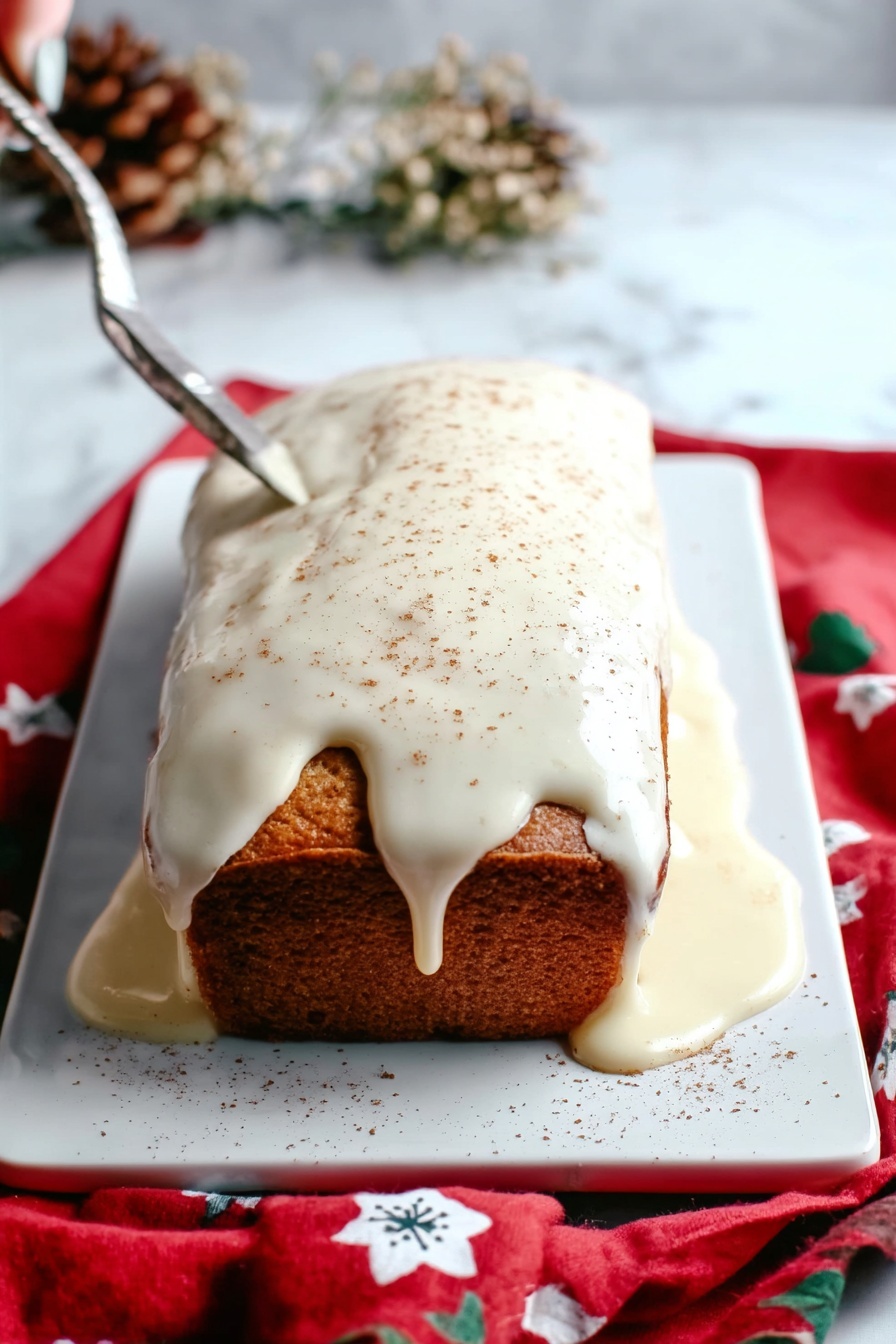 Eggnog Bread Holiday Bread Festive Eggnog Bread Christmas Bread Easy Eggnog Bread - A rectangular loaf cake sits on a white rectangular tray over a red cloth with white and blue flower patterns, placed on a white marbled surface. The cake has a golden-brown color and is covered on top with thick, creamy white icing that drips over the edges and pools around the base. The icing is lightly sprinkled with a fine brown powder, likely cinnamon or nutmeg. The scene includes dried flowers blurred in the background and a woman's hand holding a silver knife positioned above the cake. Photo taken with an iphone --ar 2:3 --v 7