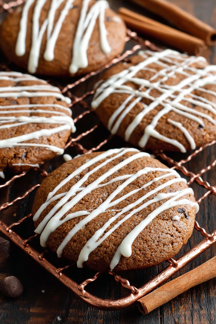 Soft Gingerbread Cookies, Gingerbread Cookies Recipe, Easy Gingerbread Cookies, Holiday Gingerbread Cookies, Chewy Gingerbread Cookies - The image shows four round brown cookies with a slightly cracked surface, placed on a copper cooling rack. Each cookie is decorated with thin, white icing drizzled randomly across the top, creating a light pattern. The cookies look soft and moist with dark spots indicating spices or chocolate chips. The background includes a dark wooden table with two cinnamon sticks placed on the right side. photo taken with an iphone --ar 2:3 --v 7