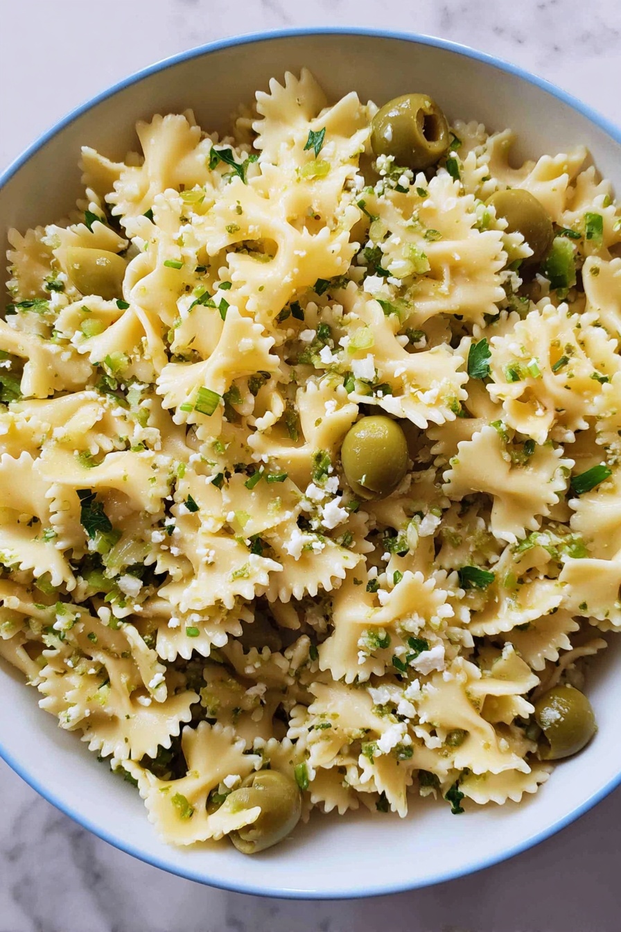 Dirty Martini Pasta Salad, savory pasta salad with olives and herbs, easy pasta salad recipes, briny pasta salad ideas, crowd-pleasing pasta salad - A close-up view of a large white bowl filled with curly-edged pasta that is light yellow in color, mixed evenly with bright green sliced olives and sprinkled with small bits of light green herbs and crumbled white cheese. The pasta is separated nicely, showing the wavy edges and soft texture, while the green olives add contrast throughout. The bowl sits on a white marbled surface, creating a clean and fresh look. photo taken with an iphone --ar 2:3 --v 7