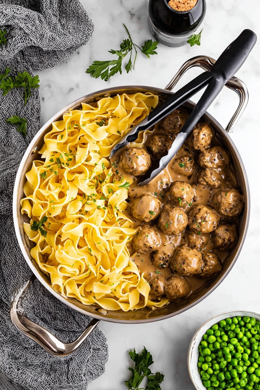 Swedish Meatballs with Creamy Gravy, Swedish Meatballs, Creamy Gravy Recipe, Comfort Food Recipes, Easy Swedish Meatballs - A large silver pan with tall handles holds two distinct layers inside: one side is filled with yellow wide pasta noodles twisted and folded over each other with a smooth, slightly shiny texture, while the other side has many round brown meatballs covered in a thick, creamy brown sauce with visible black pepper specks. Metal tongs with black grips rest on top of the pasta, slightly lifting some noodles. The pan is placed on a white marbled surface, surrounded by fresh green parsley leaves scattered around, a gray textured cloth to the left, a white bowl filled with bright green peas on the bottom right, and a dark bottle with cork inside at the top. Photo taken with an iphone --ar 2:3 --v 7