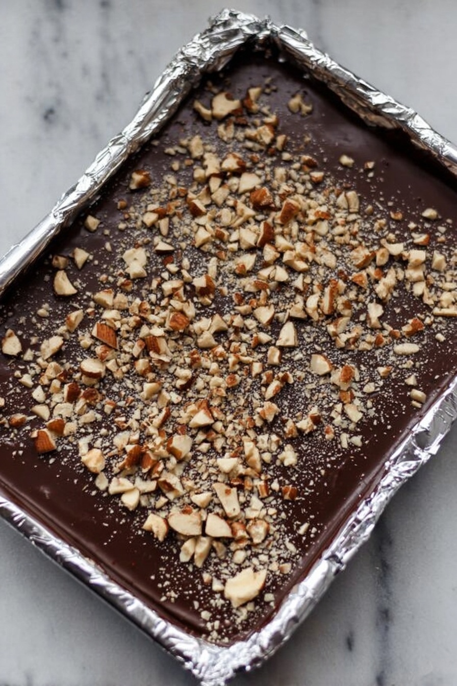English Toffee with Chocolate and Nuts, easy toffee recipe, homemade toffee bites, crunchy toffee with chocolate, nutty caramel toffee - This image shows a foil-lined rectangular tray filled with a smooth, dark chocolate layer. On top of the chocolate is an even spread of chopped nuts, both larger and smaller pieces, creating a textured surface. The foil edges around the tray are crinkled and shiny, reflecting light softly. The background is a white marbled texture. photo taken with an iphone --ar 2:3 --v 7