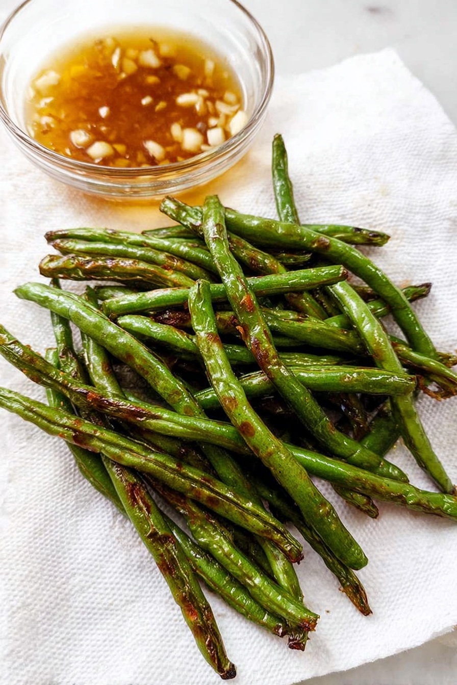 Garlic Green Beans Stir Fry, healthy vegetable side, quick green bean recipes, easy stir fry ideas, garlic vegetable stir fry - A close-up of cooked green beans piled loosely on a white plate, with each bean showing a shiny, slightly wrinkled green surface and some small pieces of browned garlic sprinkled on and around them. The beans have a mix of bright green and darker areas from cooking, and some beans are curved while others lay straight, creating a natural, uneven texture. The background is a white marbled texture. photo taken with an iphone --ar 2:3 --v 7