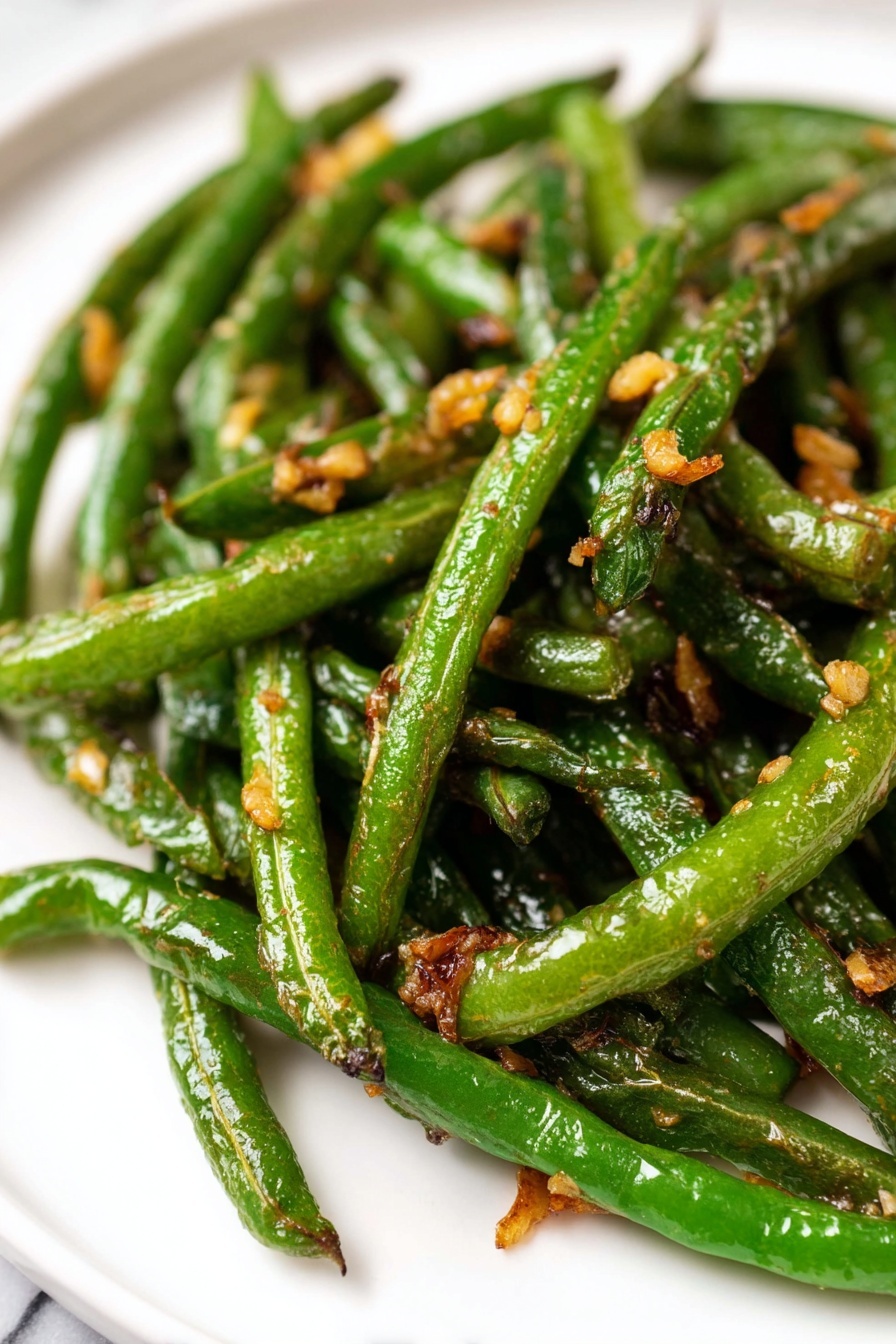 Garlic Green Beans Stir Fry, healthy vegetable side, quick green bean recipes, easy stir fry ideas, garlic vegetable stir fry - A pile of cooked green beans with browned, slightly wrinkled skin is placed on a white paper towel over a white marbled surface. To the upper left of the green beans, there is a clear round glass bowl filled with a light brown sauce mixed with small pieces of chopped garlic or onion. The sauce has a glossy texture. The background is a white cloth that partially covers the surface. photo taken with an iphone --ar 2:3 --v 7