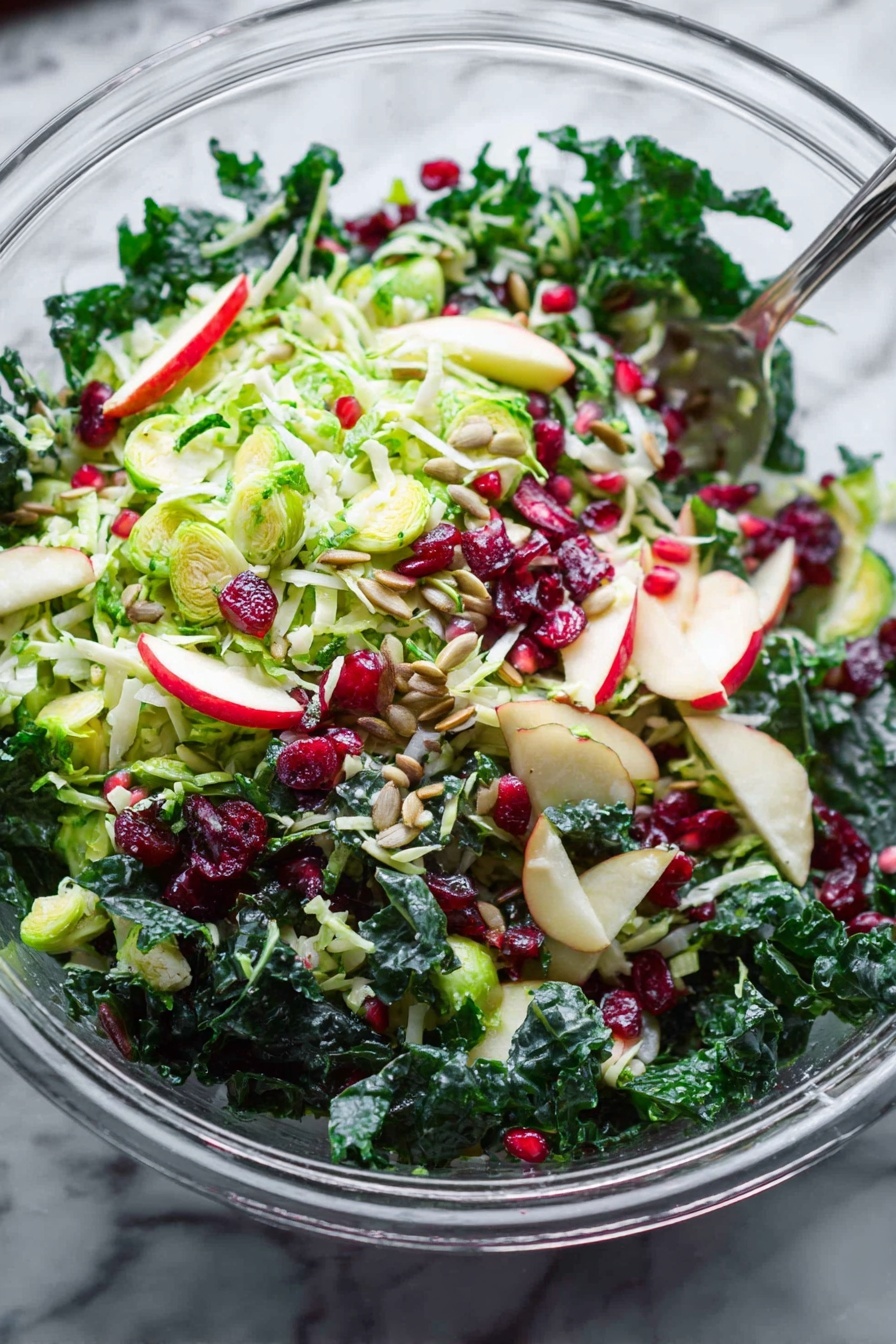 Shaved Brussels Sprouts Salad with Pomegranate, Brussels Sprouts Salad, Pomegranate Salad Recipe, Healthy Brussels Sprouts Salad, Vibrant Vegetable Salad - A clear glass bowl filled with a fresh salad on a white marbled surface, showing multiple layers of ingredients. The base layer includes dark green leafy kale with a thick texture. On top, there is a layer of thinly sliced light green Brussels sprouts mixed with small slices of light red and white apple. Small pieces of red onion add specks of purple color throughout. Scattered across the salad are bright red dried cranberries and small shiny red pomegranate seeds. Light brown sunflower seeds are spread over the salad, adding texture. A silver serving spoon is partially visible inside the bowl. The whole scene is bright and fresh, with natural light highlighting the crispness of the vegetables and fruits. Photo taken with an iphone --ar 2:3 --v 7