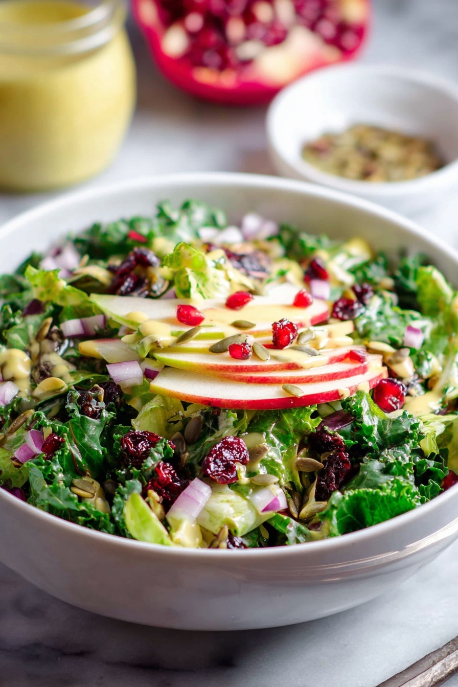 Shaved Brussels Sprouts Salad with Pomegranate, Brussels Sprouts Salad, Pomegranate Salad Recipe, Healthy Brussels Sprouts Salad, Vibrant Vegetable Salad - A white bowl is filled with a fresh salad showing multiple layers: at the bottom are bright green leafy vegetables, followed by thin slices of red and green apple. On top, there are pieces of chopped red onion, dark green kale leaves, bright red pomegranate seeds, dried red cranberries, and scattered sunflower seeds, all mixed together. The salad is lightly dressed with a pale yellow dressing that adds a slight shine to the ingredients. The bowl is placed on a white marbled surface, with a blurred jar of light yellow dressing and a small white bowl of seeds in the background. photo taken with an iphone --ar 2:3 --v 7