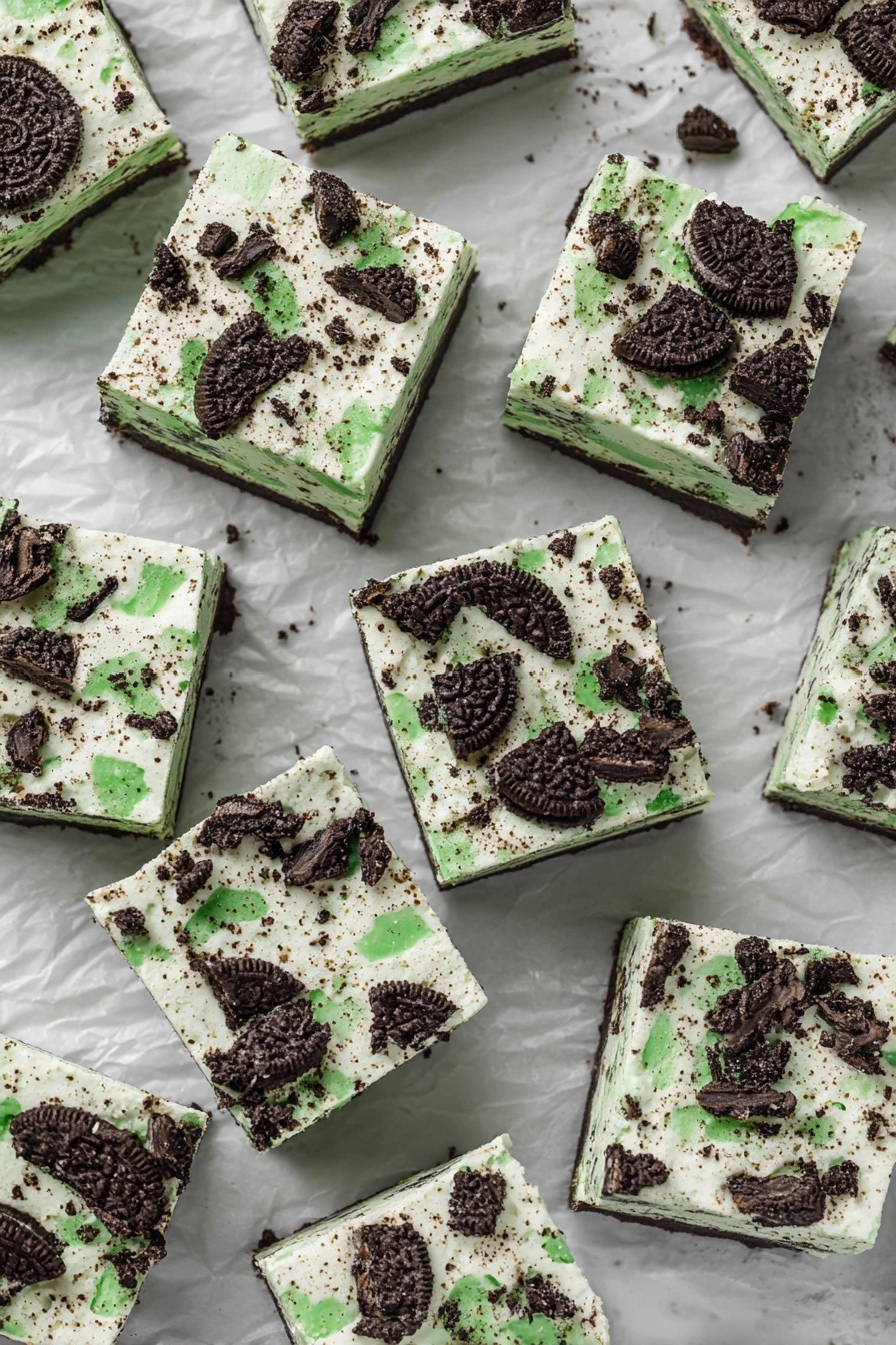 Mint Oreo White Chocolate Fudge, Mint Oreo Fudge, White Chocolate Oreo Candy, Easy Mint Fudge, No-Bake Oreo Fudge - The image shows many square pieces of dessert laid out on crumpled white paper over a white marbled surface. Each square has two visible layers: the bottom layer is a light creamy color with specks inside, and the top layer is a white cream with crushed dark chocolate cookies that have bright green filling scattered unevenly across the surface. The cookies add a rough texture on top, with pieces varying in size and shape. The squares are arranged closely but not touching each other, filling the image space evenly, and giving a look of a cool, creamy treat with a minty cookie crunch on top photo taken with an iphone --ar 2:3 --v 7