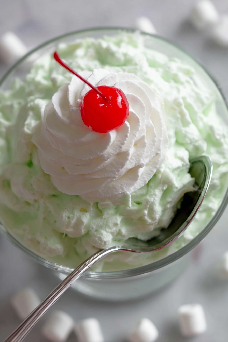 Easy Watergate Salad, Watergate Salad with Pineapple and Marshmallows, Pineapple Marshmallow Salad, Retro Watergate Salad, Simple Party Salad - A close-up image of a layered dessert in a clear glass bowl placed on a white marbled surface with mini white marshmallows scattered around. The bottom layer is a creamy, light green textured mixture that looks fluffy and thick. On top of this green layer is a smooth, white dollop of whipped cream shaped in a spiral pattern. Sitting right in the center of the whipped cream is a bright red, shiny cherry. A silver spoon is partly inserted into the green layer, curving around the side of the bowl. photo taken with an iphone --ar 2:3 --v 7