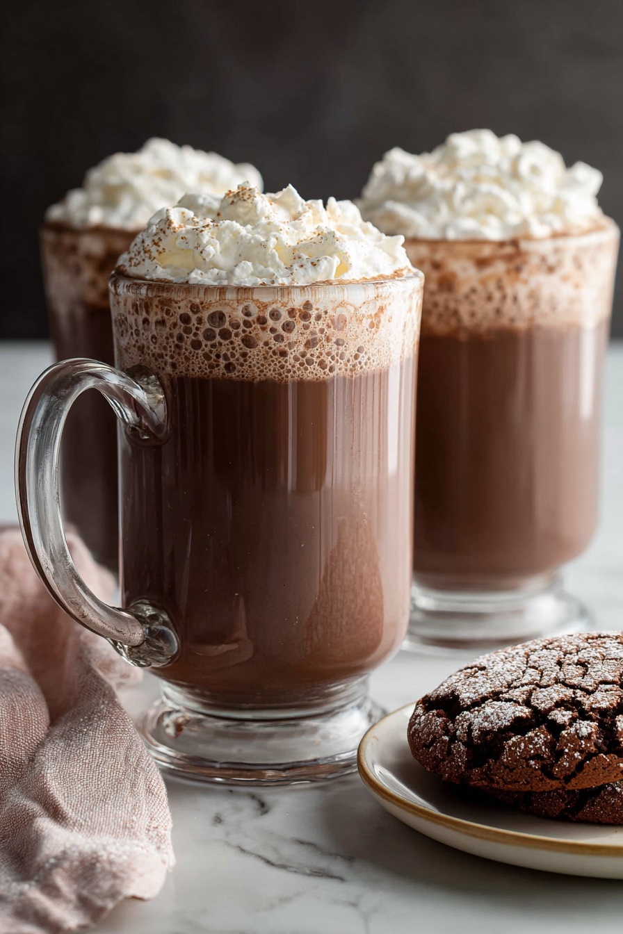 Homemade Hot Chocolate, easy hot chocolate recipe, creamy hot chocolate, rich hot chocolate drink, winter warm-up recipes - Two clear glass mugs filled with rich brown hot chocolate, topped with a thick layer of light white whipped cream that has a fluffy texture and soft peaks. The mugs have sturdy handles positioned on the left, and the hot chocolate shows slight bubbles near the edges under the whipped cream. In the foreground, on the right side, there is a white plate with a beige rim holding two cracked chocolate cookies with a powdery surface. A soft pink and white cloth napkin is casually placed on the left side near the front mug. All items sit on a smooth white marbled surface with a dark, blurred background behind them. Photo taken with an iphone --ar 2:3 --v 7